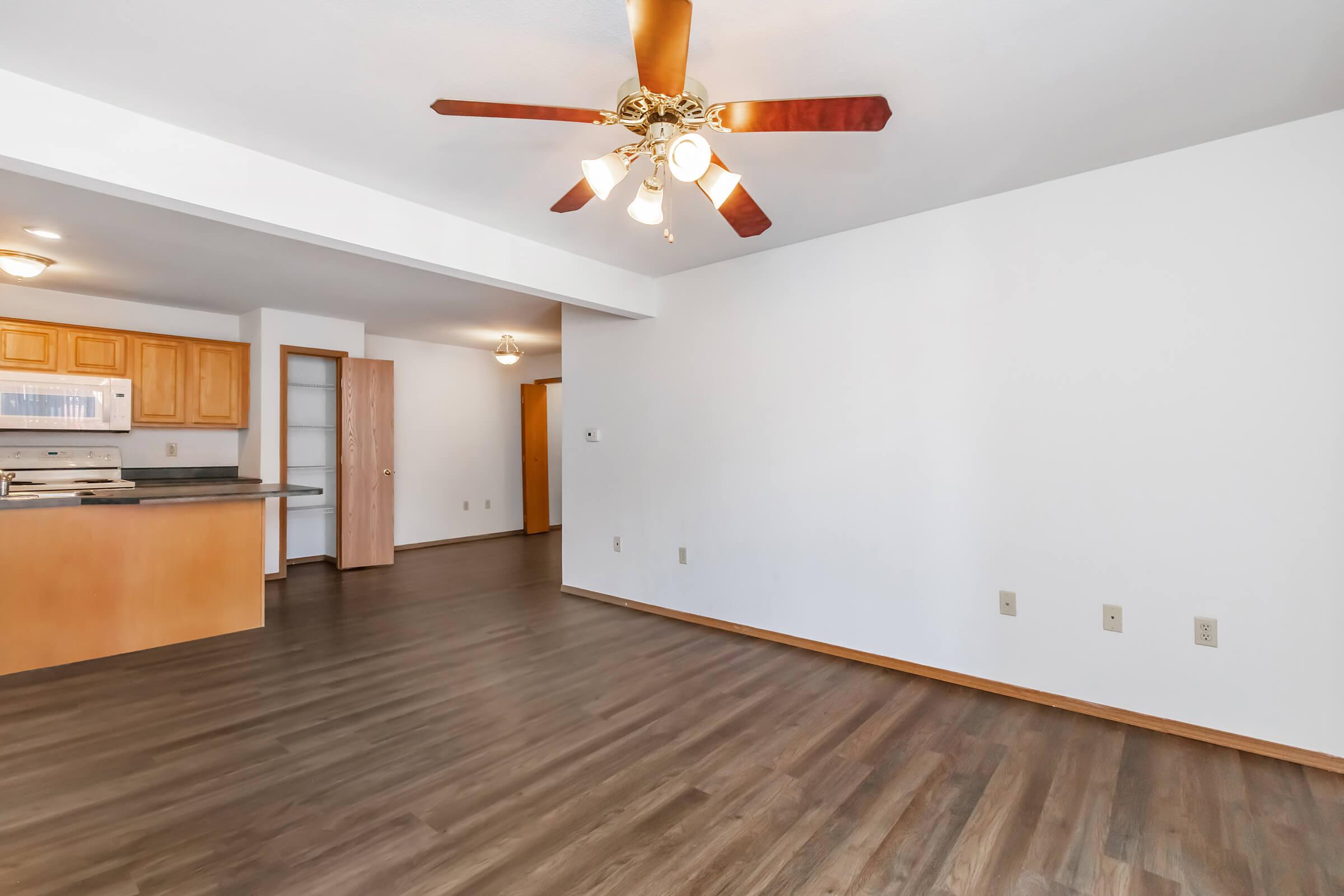 Spacious living area with a ceiling fan, featuring hardwood floors and neutral-colored walls. A kitchen with wooden cabinets and a breakfast bar is visible in the background, along with a doorway leading to another room. Natural light fills the space, creating an inviting atmosphere.