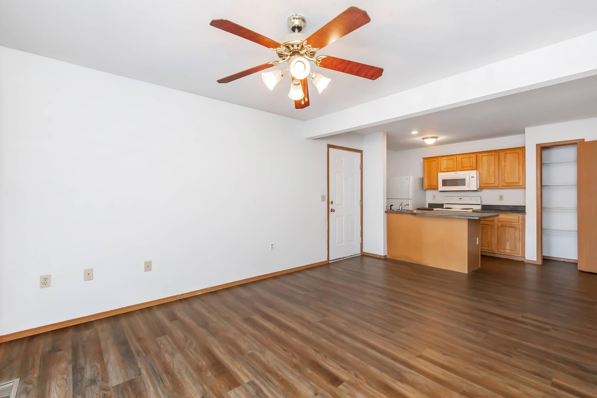A spacious living area featuring a ceiling fan with light fixtures, hardwood-style flooring, and white walls. In the background, a kitchen with wooden cabinets and appliances is visible. The layout is open and inviting, with natural light illuminating the room.