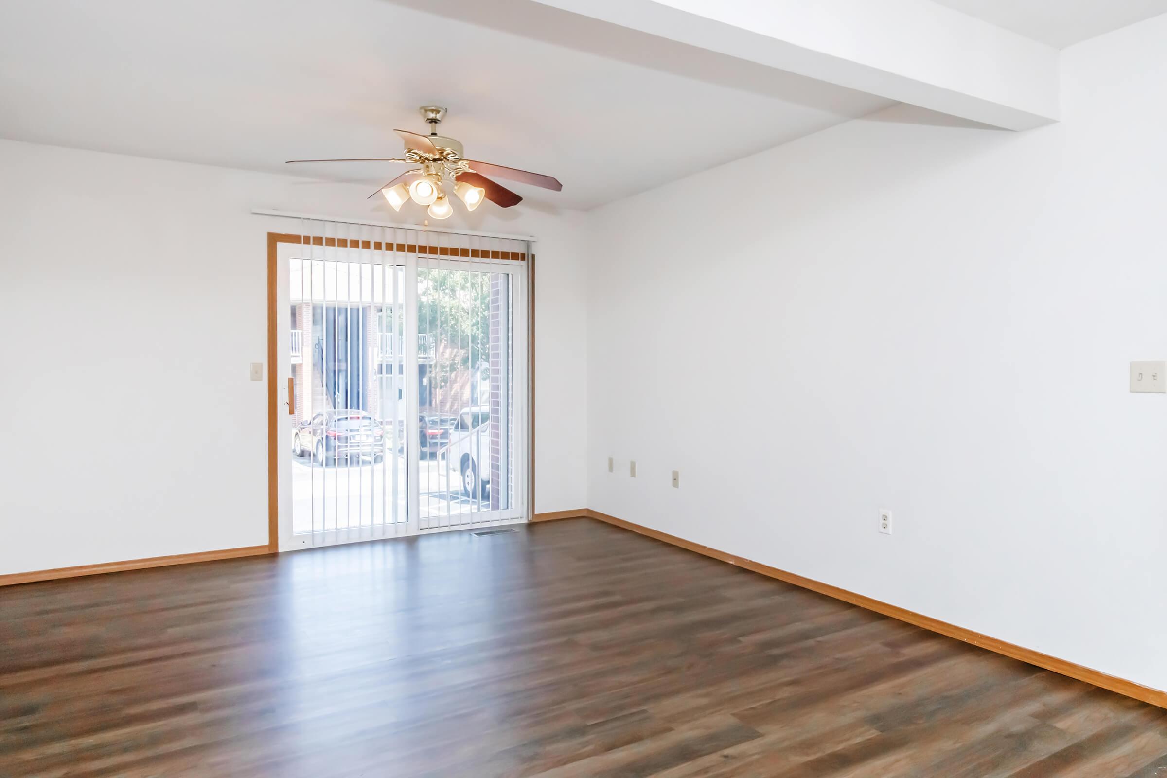Empty living room with wooden flooring, a ceiling fan with light fixtures, and a sliding glass door leading to an outdoor area. The walls are painted white, and there are no furnishings, creating a spacious and minimalistic appearance.