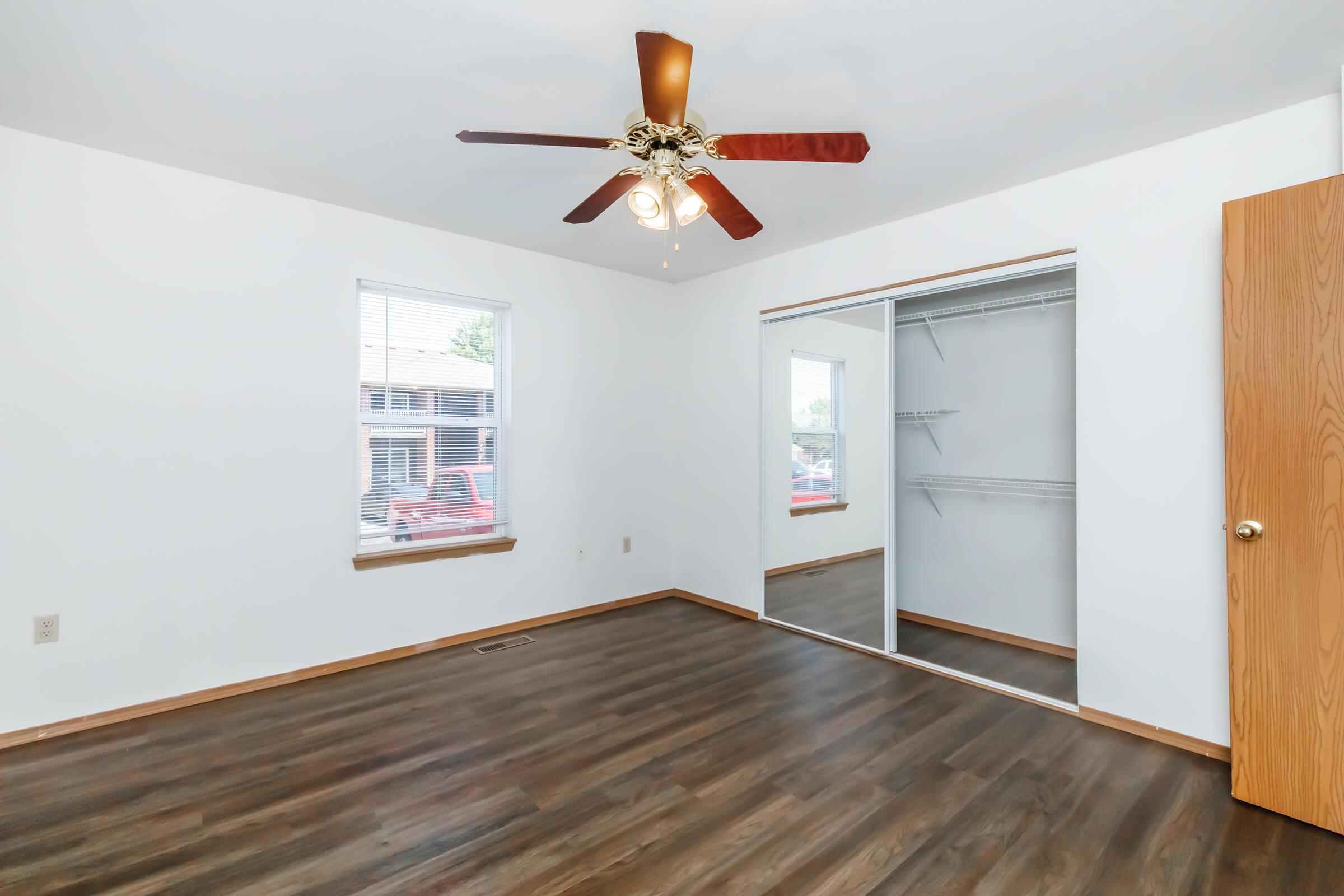 A bright, empty bedroom featuring wooden flooring, a ceiling fan with light, and two windows. There is a mirrored closet sliding door on one wall and a wooden door leading to the hallway. The walls are painted white, creating a spacious and airy feel.