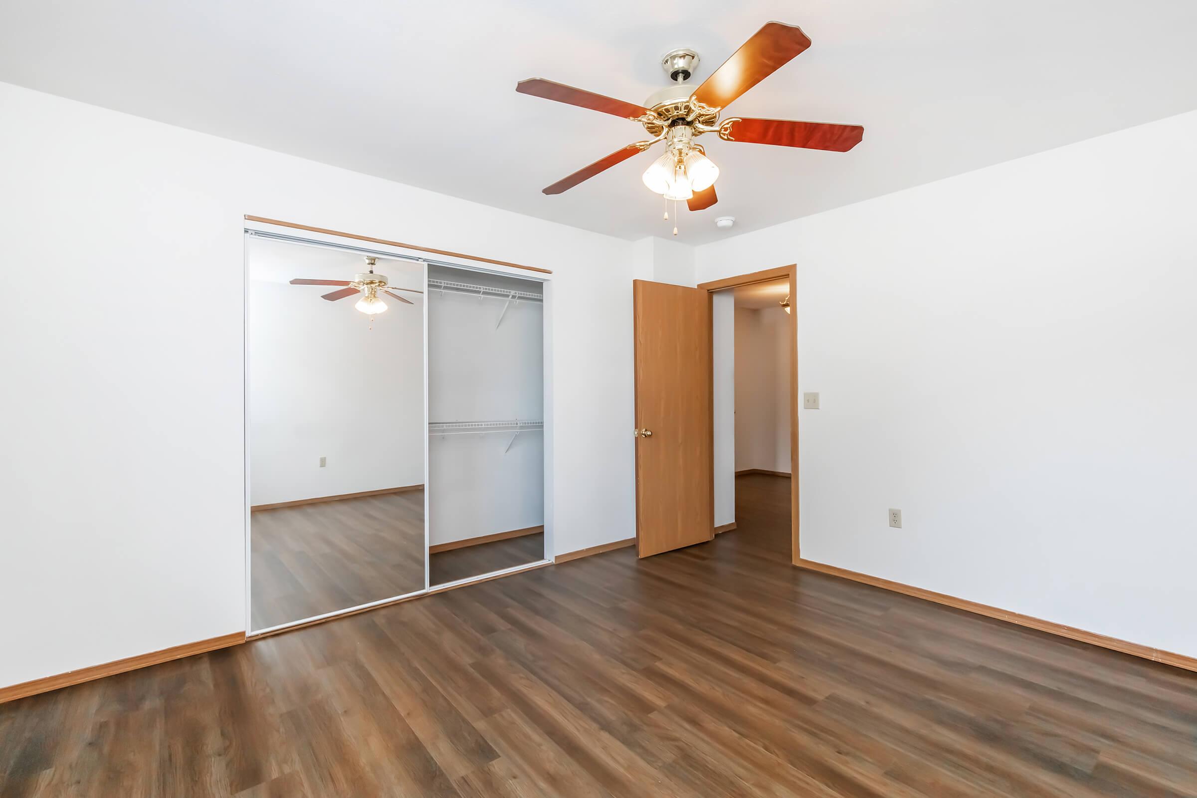 Bright, empty bedroom featuring a ceiling fan with wooden blades, light fixture, and mirrored sliding closet doors. The room has light-colored walls and a warm wood laminate floor, creating a spacious and inviting atmosphere. A wooden door leads to another area, enhancing the open layout.