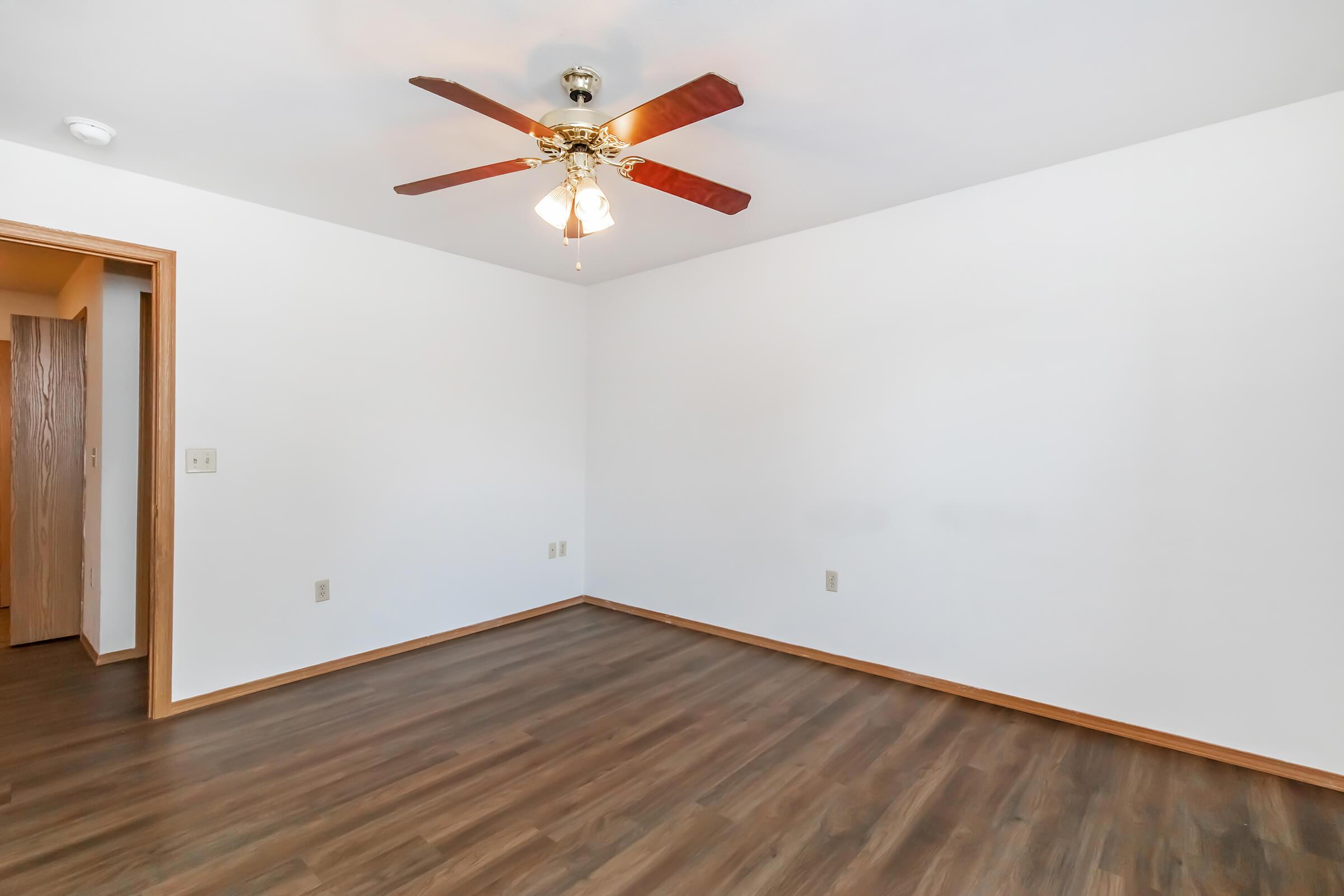 Empty room with light brown hardwood flooring and white walls. A ceiling fan with light fixtures is visible in the center, and there is an open door to another room on the left. The space is well-lit and uncluttered, creating a clean and neutral atmosphere suitable for various purposes.