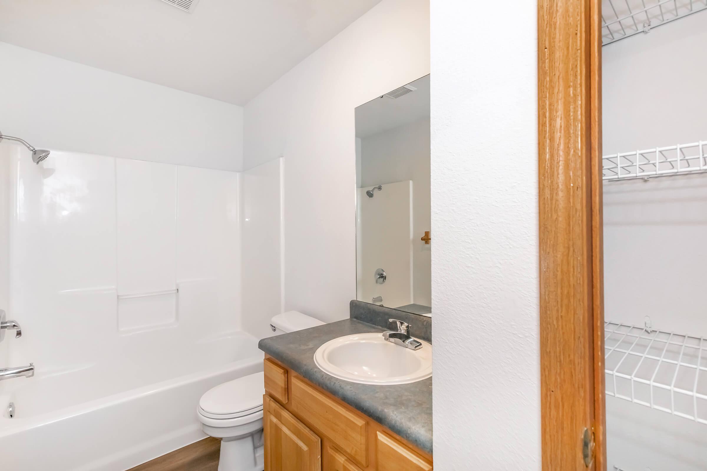A clean, modern bathroom featuring a white tub and shower enclosure, a sink with wooden cabinetry, a large mirror, and a wire shelving unit visible in the corner. The walls are painted light, contributing to a bright and spacious feel.