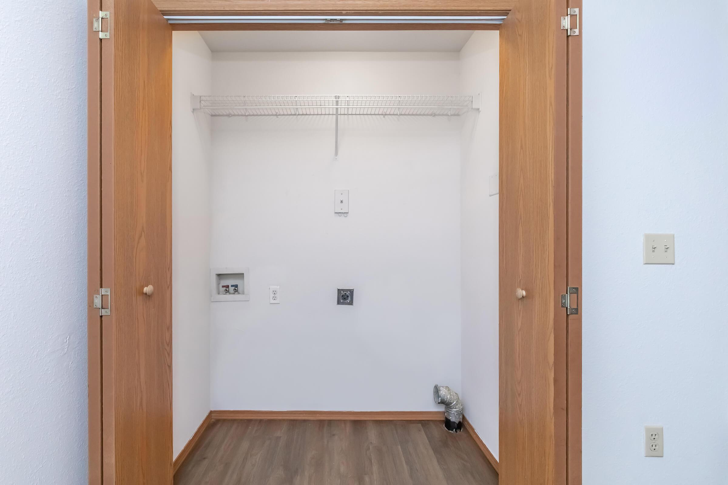 An empty closet with double wooden doors, featuring a white interior and a shelf along the back wall. There are electrical outlets and a cable connection visible on the side walls, and the floor is finished with light-colored wood laminate.