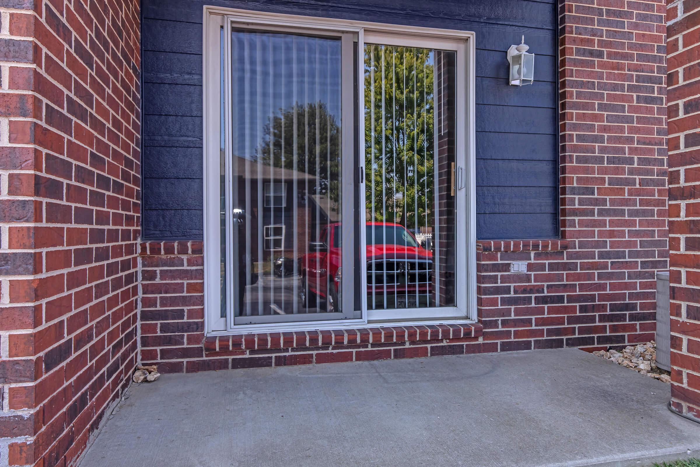 A sliding glass door with vertical blinds, situated in a brick-walled exterior. The door reflects trees and a red vehicle outside, with a simple concrete porch in front. A light fixture is mounted beside the door.