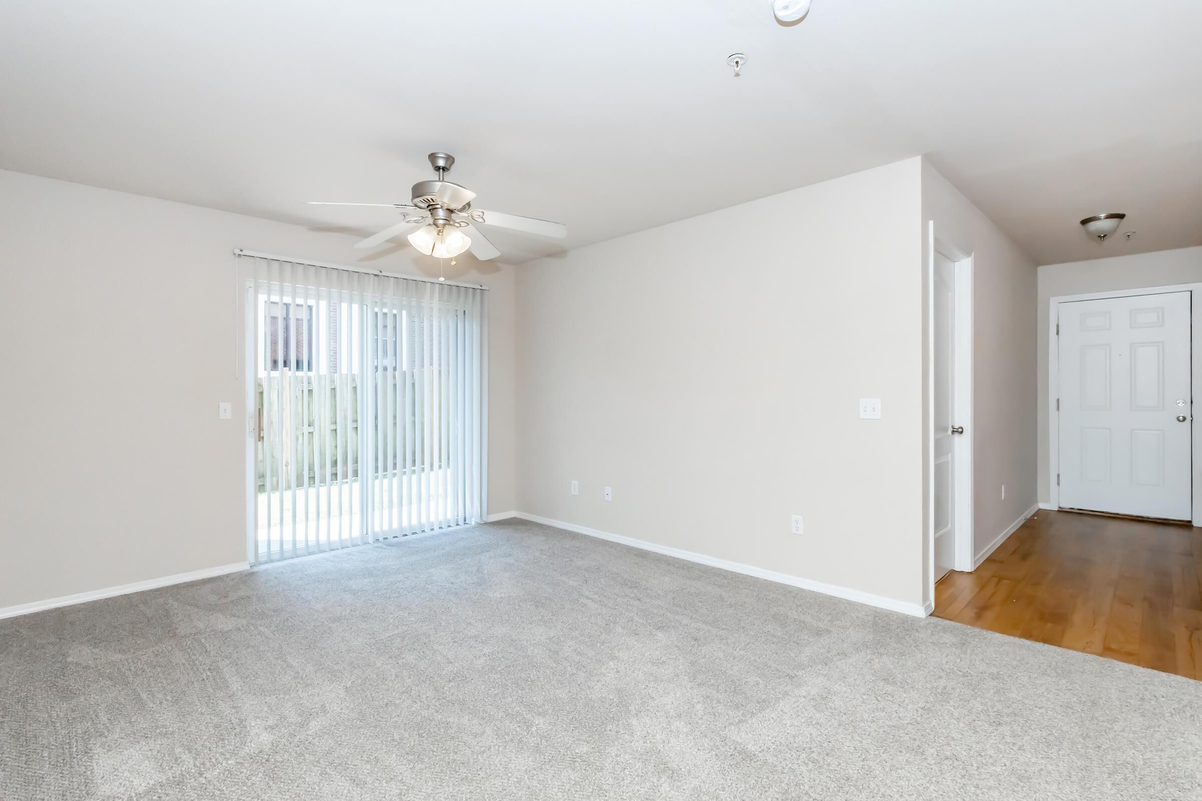 A spacious, empty living room featuring light beige walls, plush gray carpet, and a ceiling fan. There are sliding glass doors leading to a private outdoor area, and a doorway on the right side visible. The overall ambiance is bright and inviting, suitable for various decor styles.