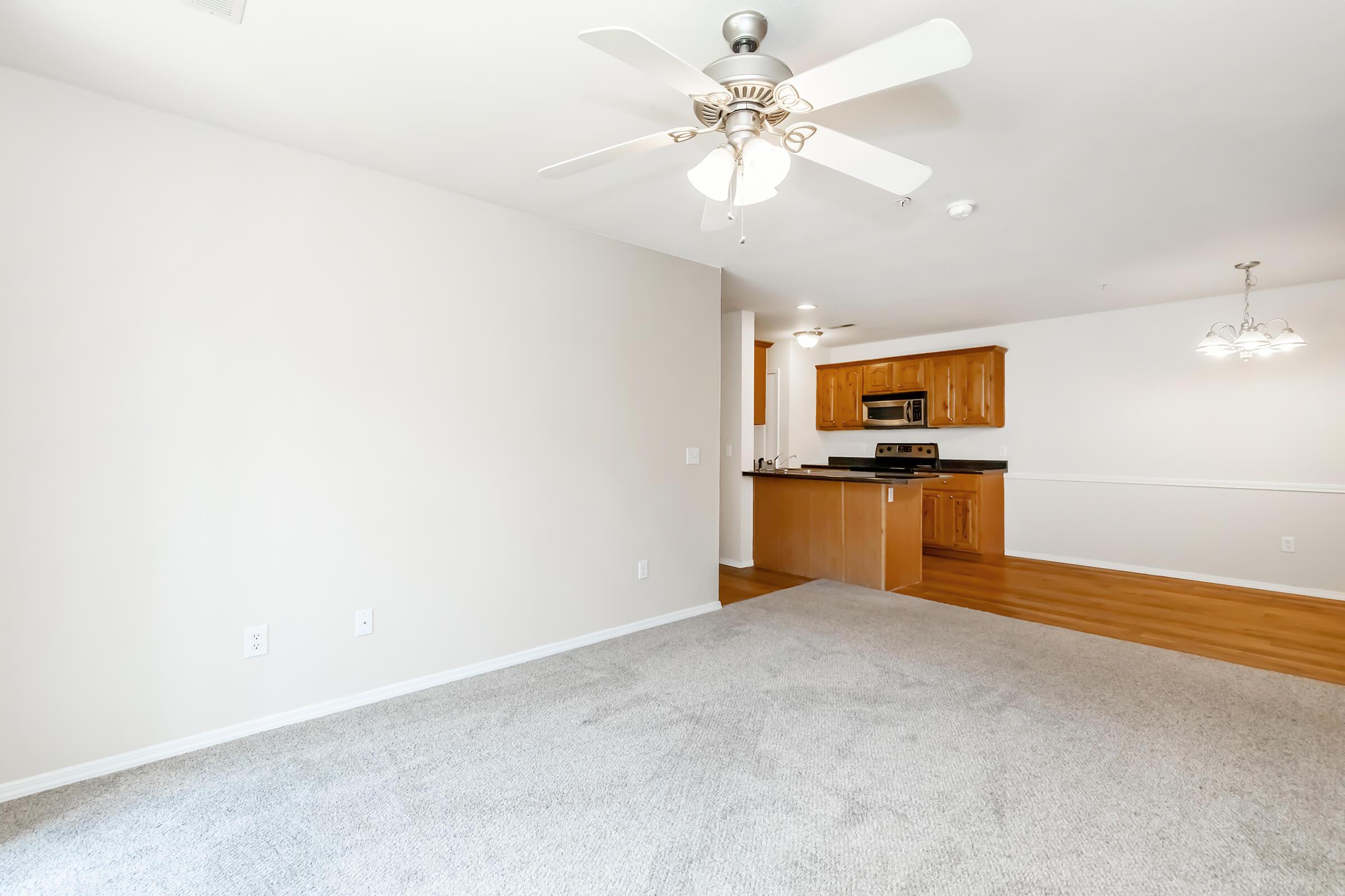 A spacious living area with light-colored carpet and a ceiling fan. In the background, there is a kitchen featuring wooden cabinets and stainless steel appliances. The walls are painted in neutral tones, providing a bright and airy atmosphere.