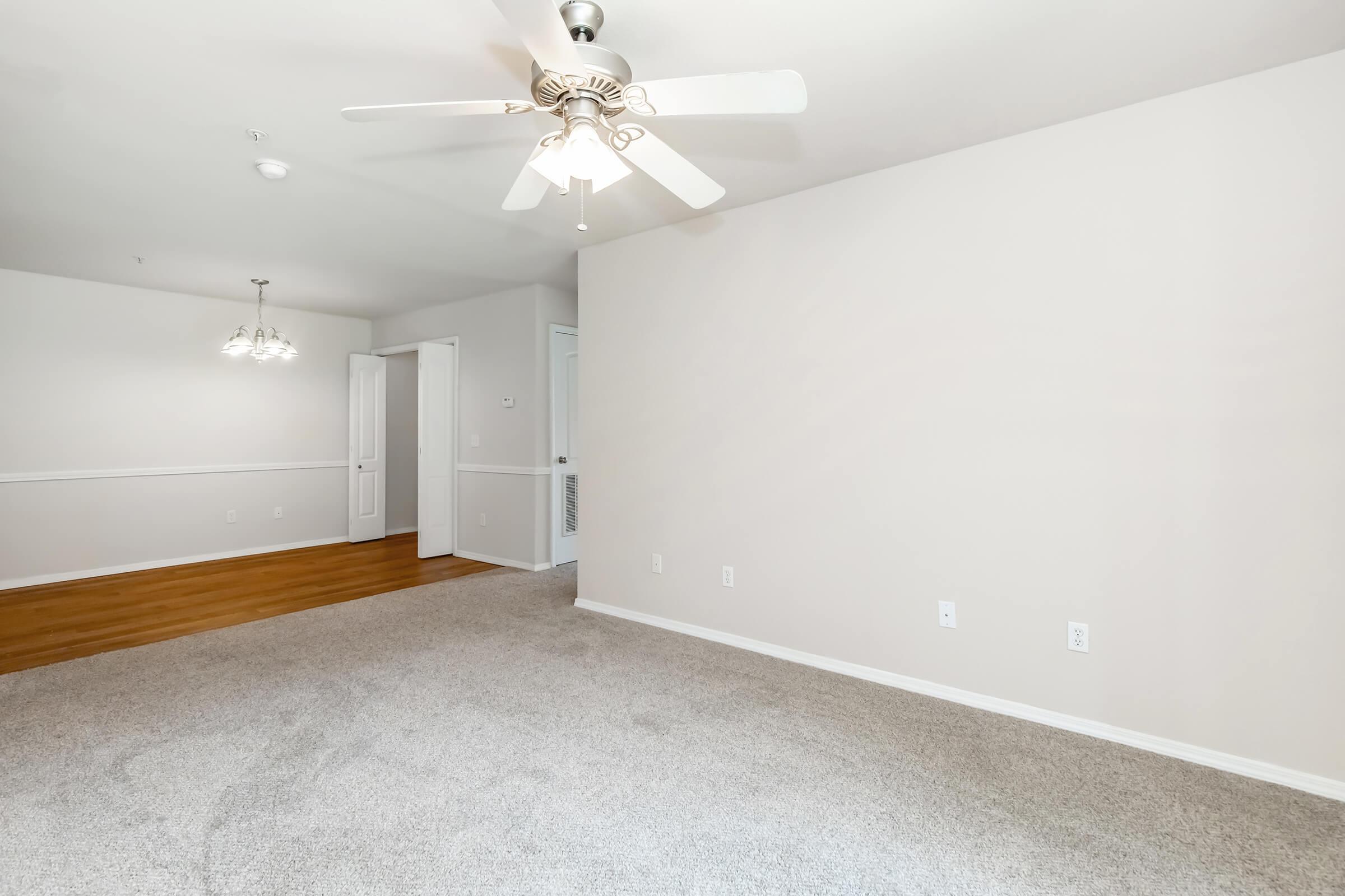 A spacious, empty living room featuring light beige walls and plush carpet. A ceiling fan hangs from the ceiling, and there is a doorway leading to another room. To the left, a small dining area is visible, adorned with two light fixtures. The overall atmosphere is bright and inviting.