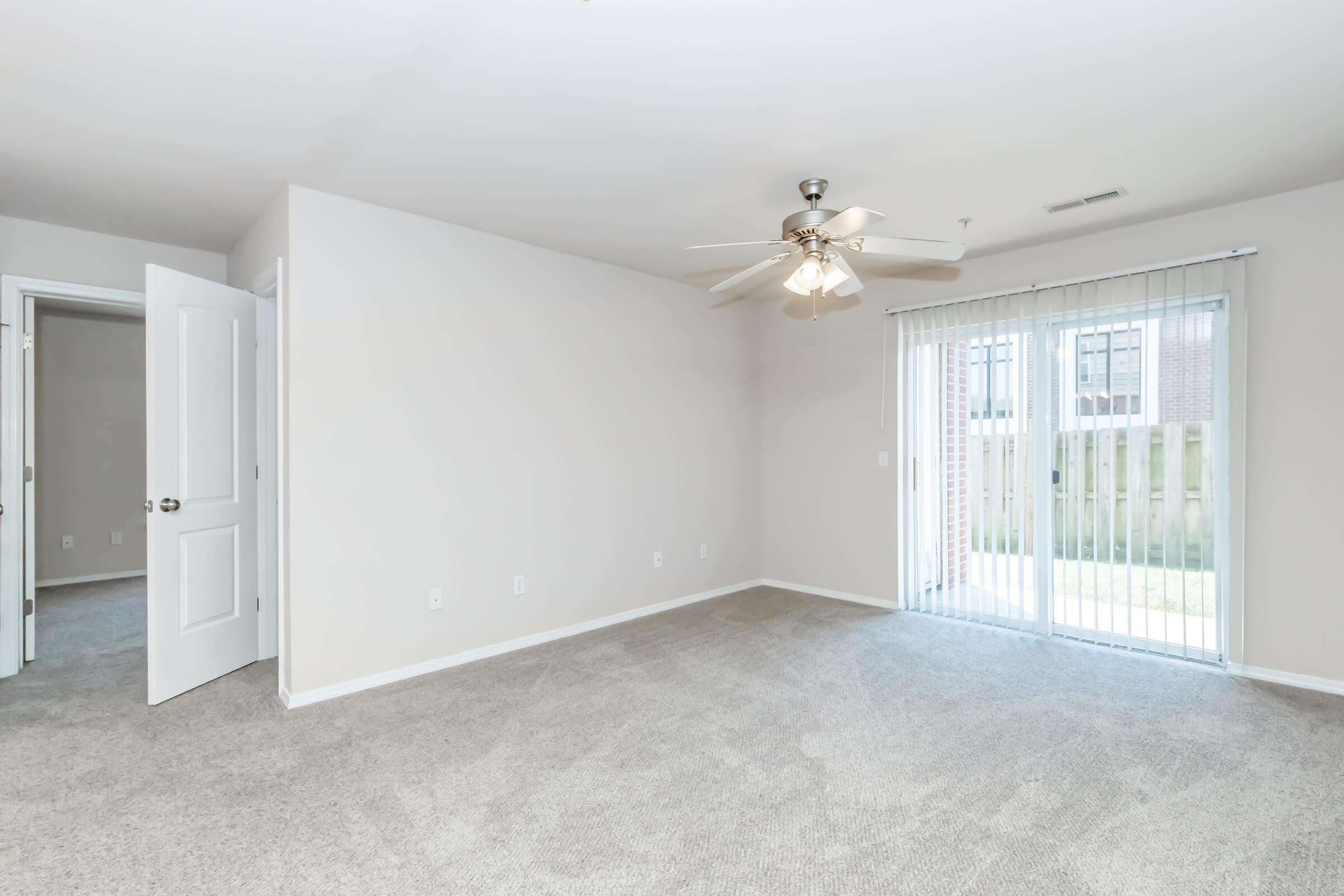 A spacious, empty room with a ceiling fan, light-colored walls, and a plush carpet. There is a sliding glass door that opens to an outdoor area, and an open doorway leads to another room. Natural light brightens the space, creating a welcoming atmosphere.
