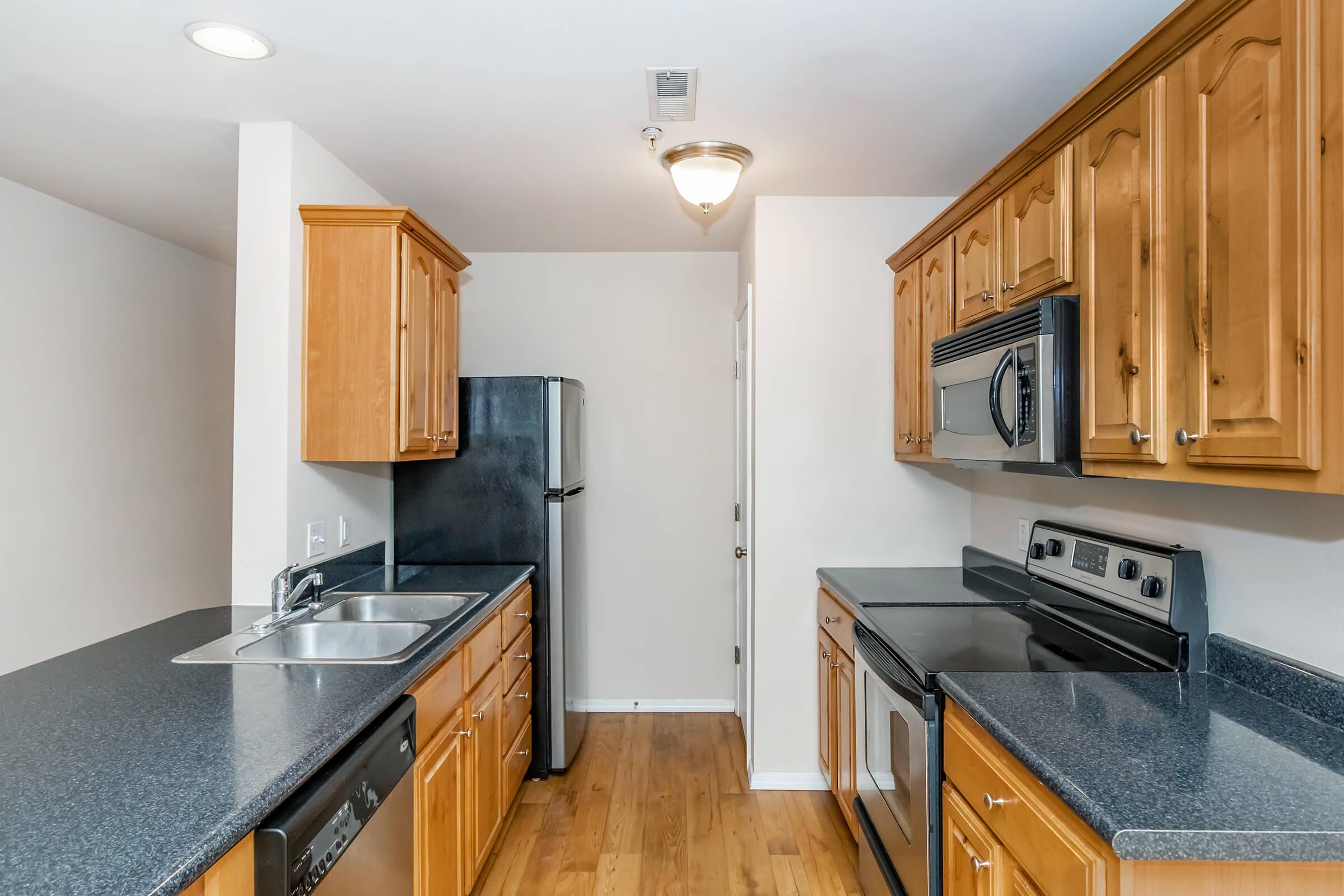 Modern kitchen featuring wooden cabinetry, black appliances, and a spacious countertop. The layout includes a sink, stove, refrigerator, and microwave, with hardwood flooring and a warm, neutral color scheme. Bright lighting enhances the inviting atmosphere.
