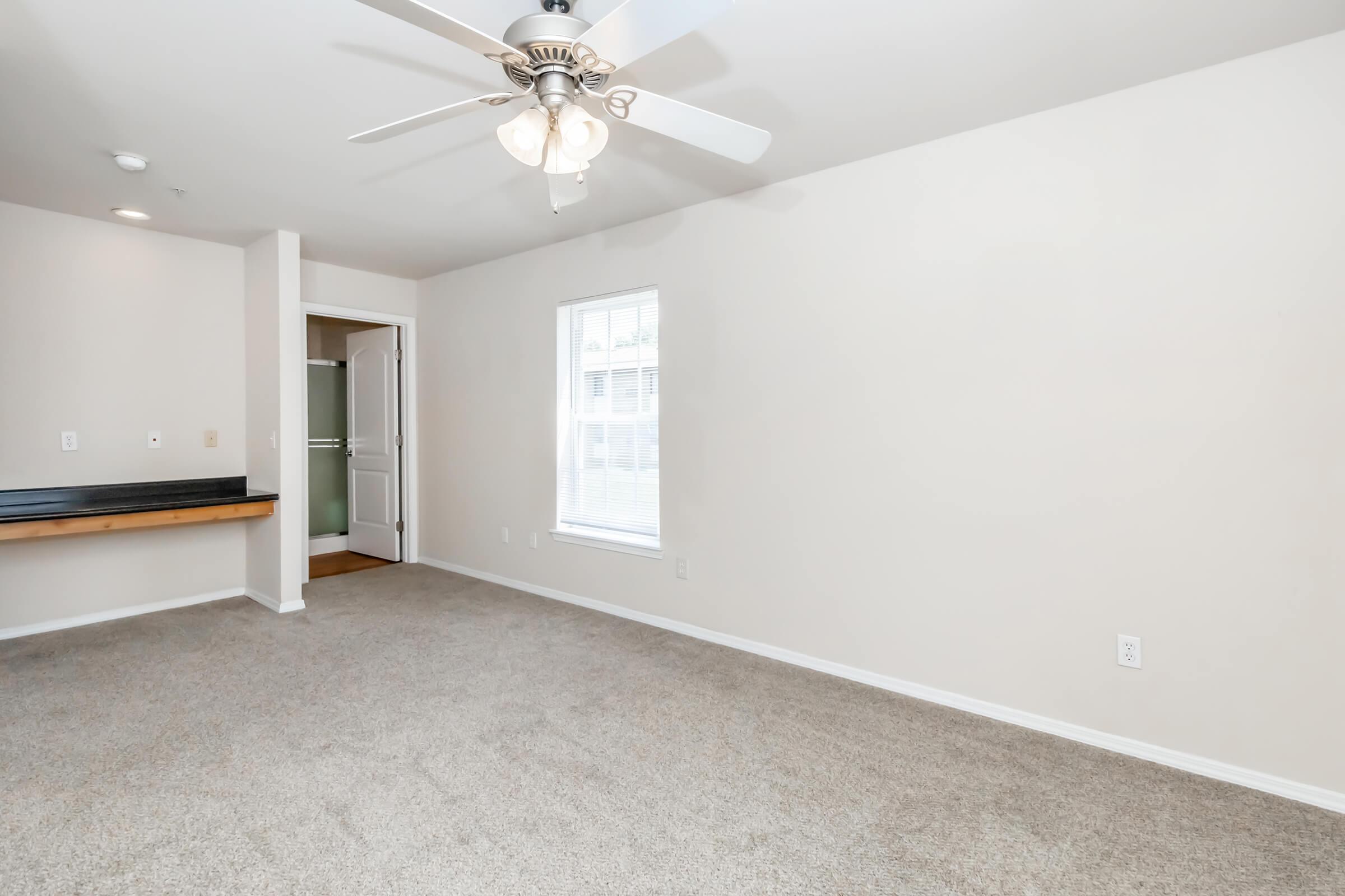 A spacious, light-colored living room with beige carpeting, featuring a ceiling fan and a large window. There is a small kitchen area visible on the left with a counter. The walls are plain and unadorned, creating a minimalist aesthetic. The room appears bright and inviting.