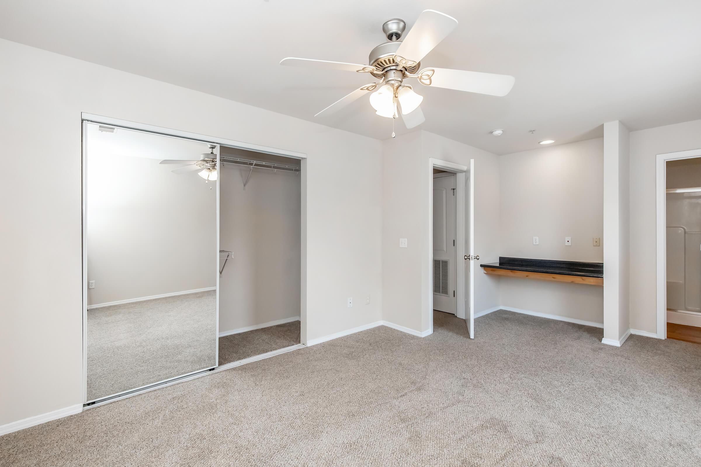 A spacious, well-lit bedroom featuring a ceiling fan, beige carpeting, and neutral-colored walls. It includes a mirrored sliding closet door, a small shelf along one wall, and an en-suite bathroom area visible through an open door. The room feels clean and inviting.