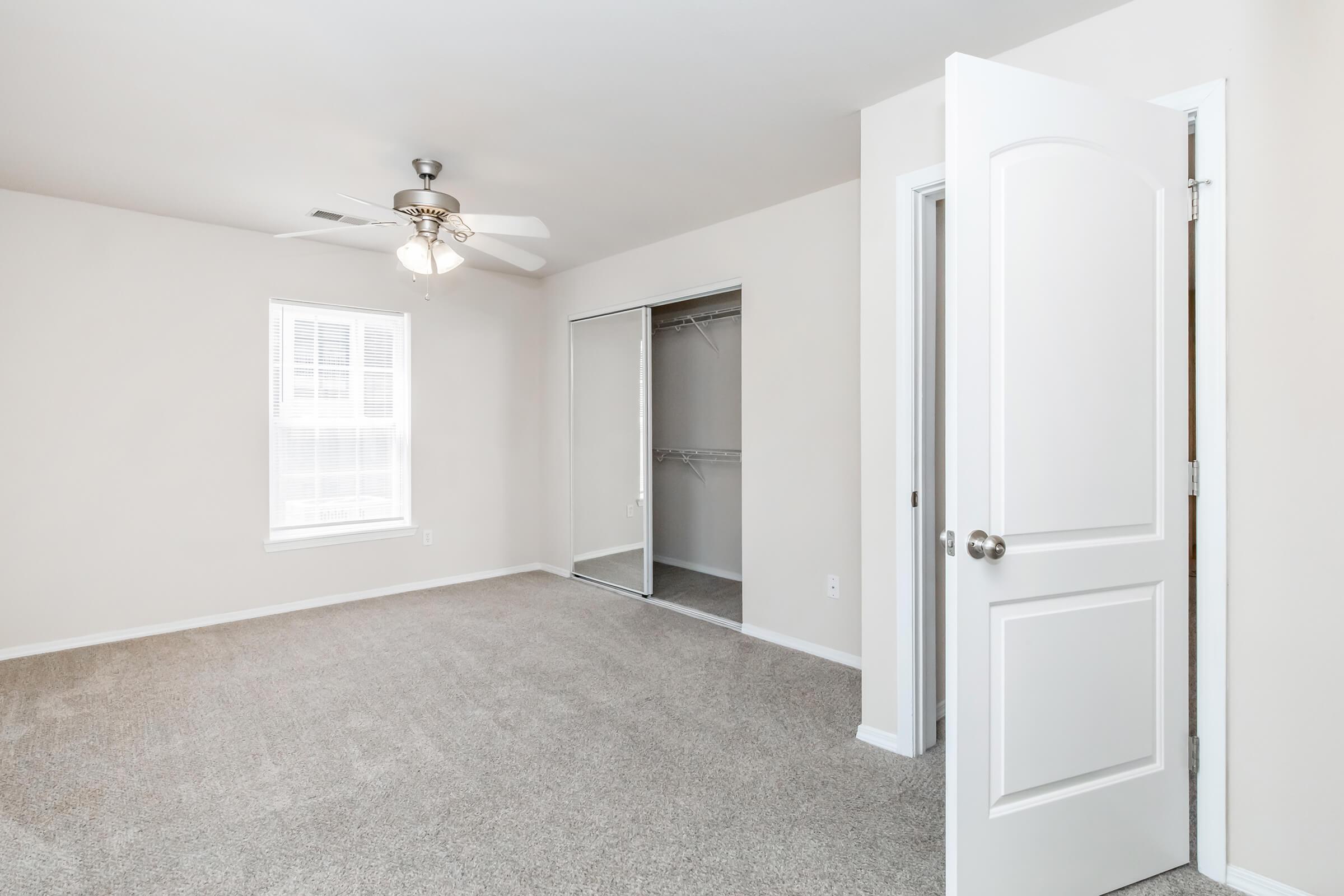 A clean, empty bedroom showcasing light-colored walls and a ceiling fan. There is a window allowing natural light, and a spacious closet with sliding doors on one side. The flooring is carpeted, creating a cozy atmosphere. The door is open, giving a view of the room's layout.