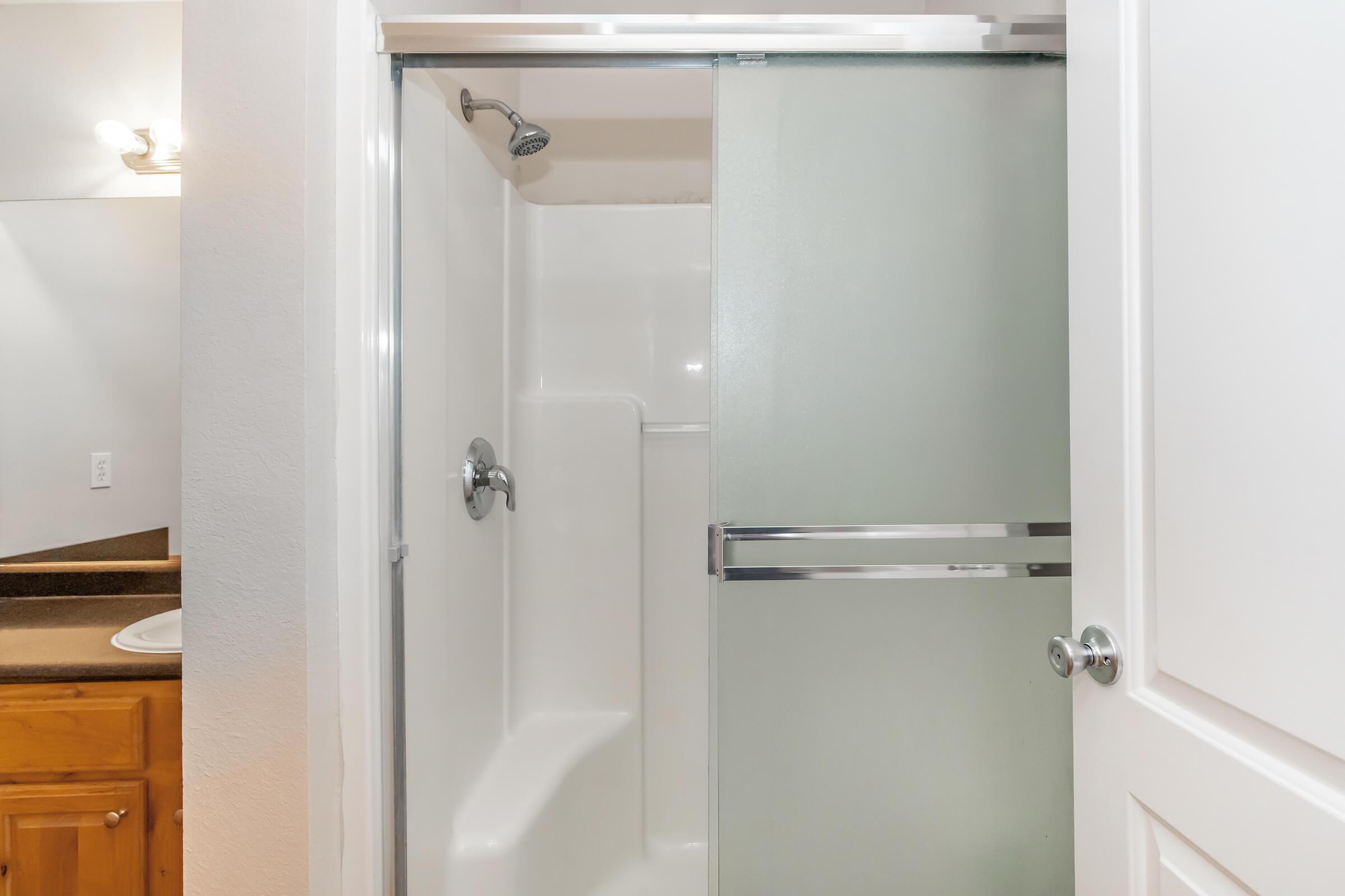 A clean bathroom featuring a walk-in shower with a glass door. The shower has a chrome faucet and a small shelf, while the surrounding area includes light-colored walls and wood cabinetry. A mirror and countertop are visible on one side. Gentle lighting creates a bright atmosphere.