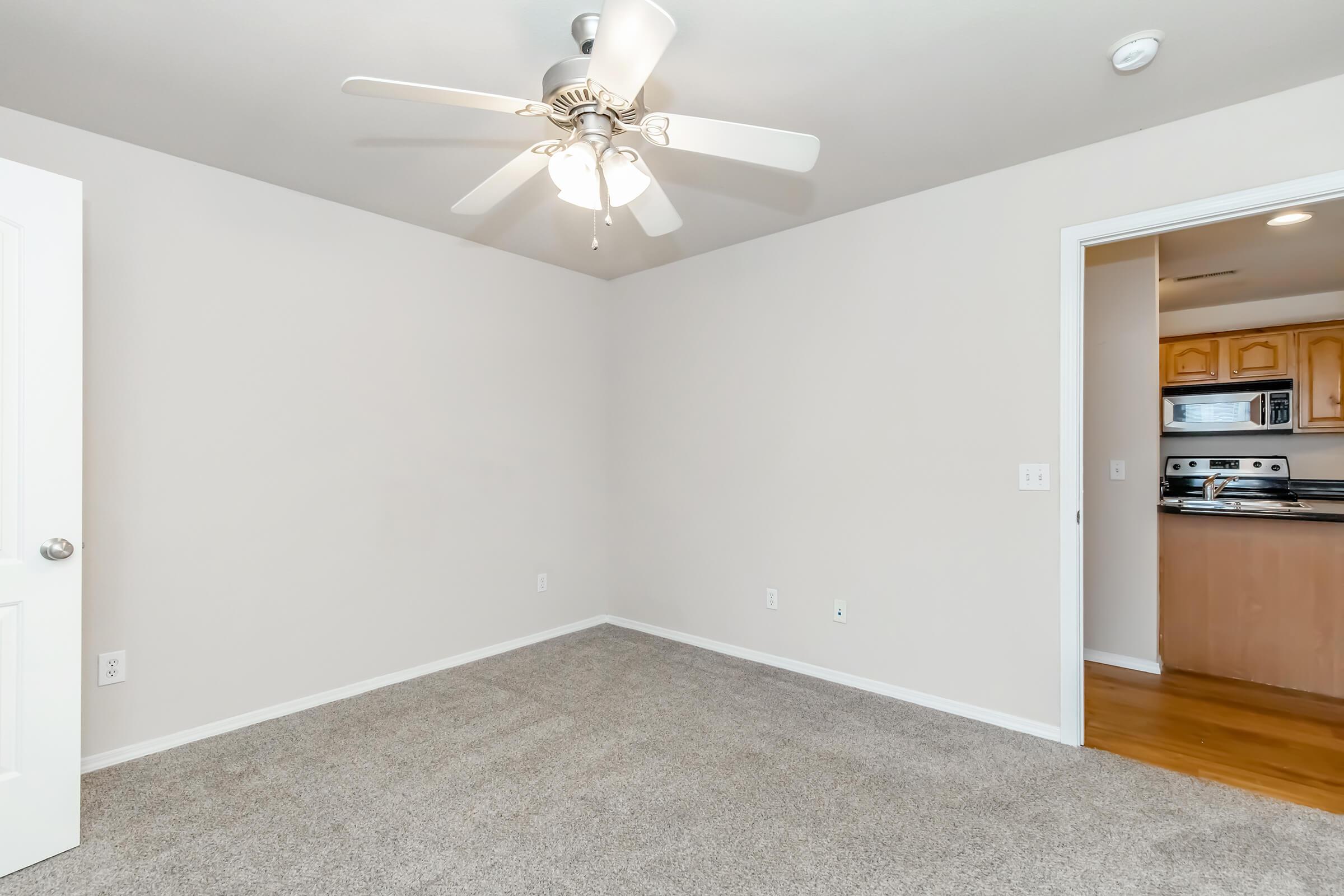 Empty room with light gray carpet flooring, a ceiling fan with light, and beige walls. A doorway on the left leads to another room with a glimpse of a kitchen. The space appears clean and spacious, suitable for various furniture arrangements.