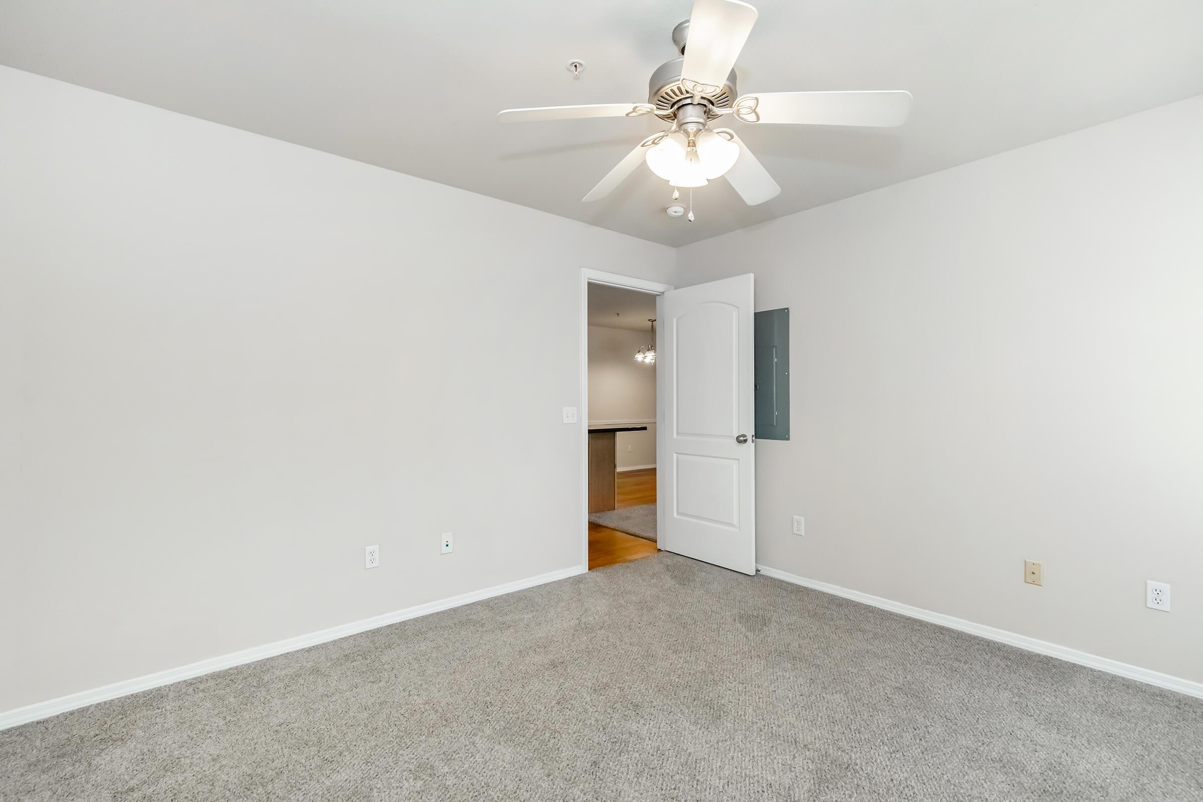 A well-lit, empty room featuring light gray carpet and a ceiling fan. There is a white door leading to another room on the left side, and the walls are painted a neutral color. The space appears clean and spacious, suitable for various uses such as a bedroom or office.