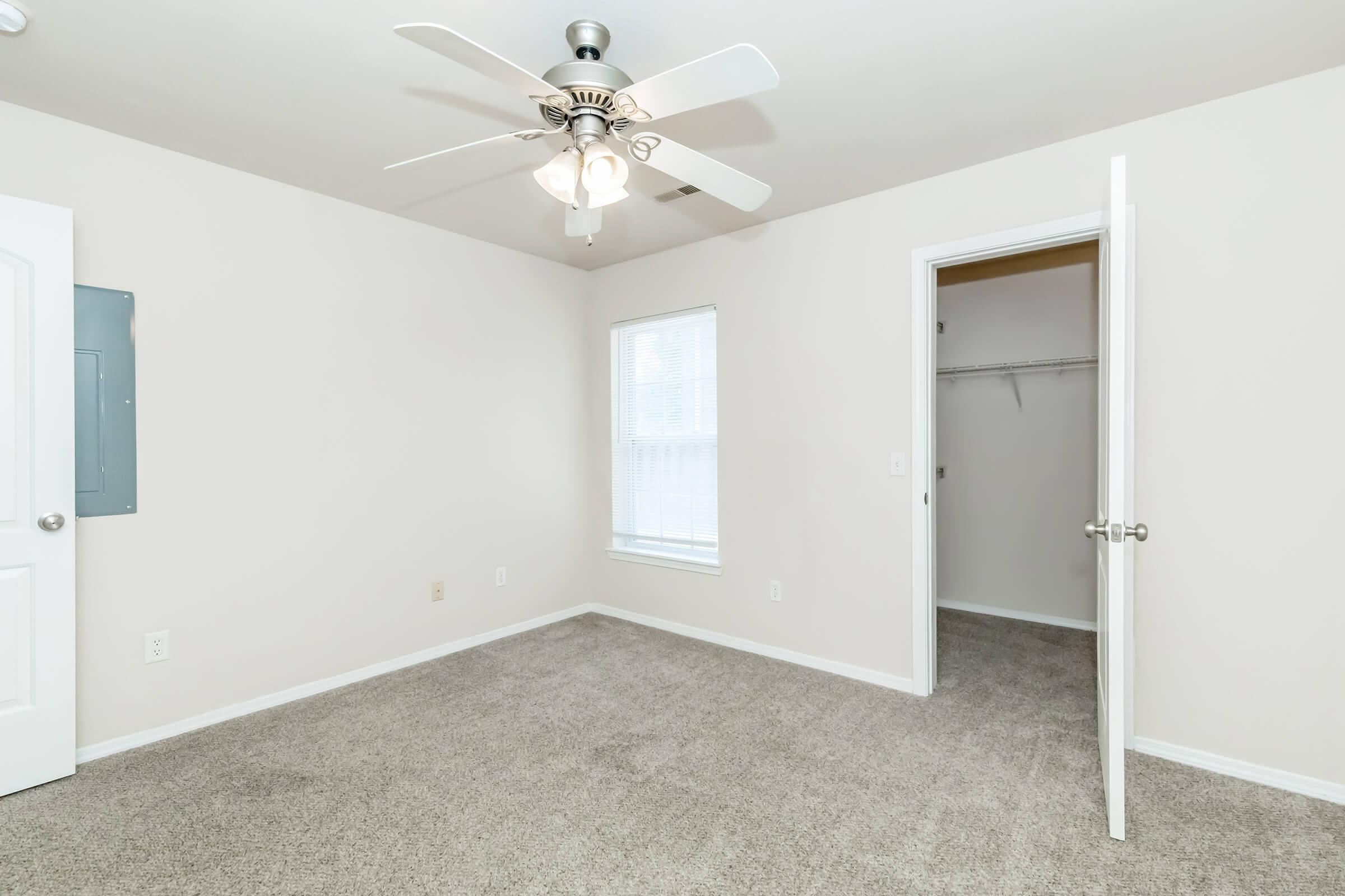 Interior view of a light-colored bedroom featuring a ceiling fan, neutral carpet, and a window letting in natural light. An open door leads to a closet on the right, while the left wall has an electrical panel. The overall ambiance is clean and simple, suitable for various decor styles.