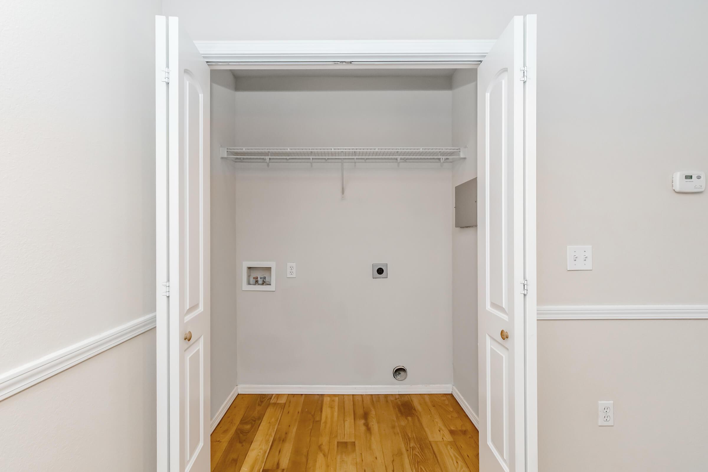 A clean, empty laundry room with white double doors open wide. The walls are light-colored, and there is a wire shelf above. The floor is wooden, and the room features a small wall-mounted panel and a circular hole in the floor, indicating plumbing or electrical connections.