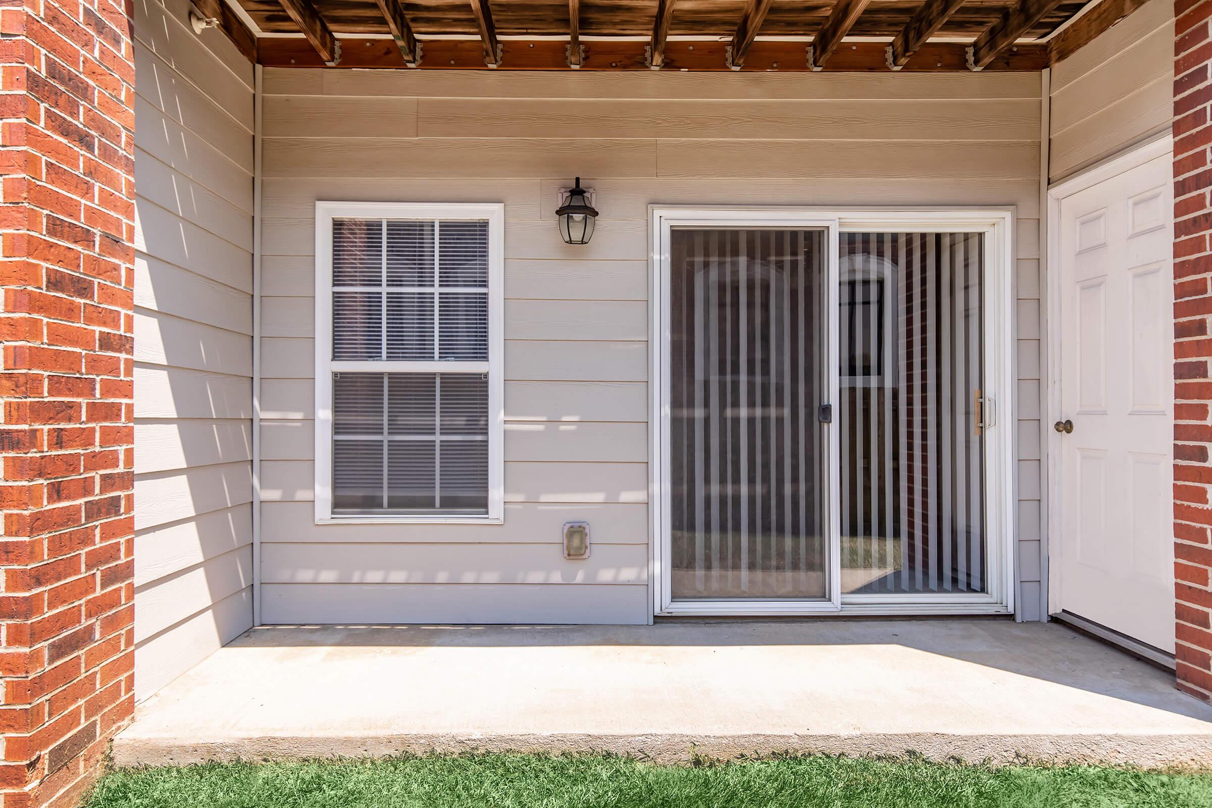 A small outdoor patio area featuring a sliding glass door with vertical blinds, a single window with grid patterns, a light fixture on the wall, and a white door on the right. The space has a concrete floor and is bordered by red brick columns, with a patch of green grass in the foreground.