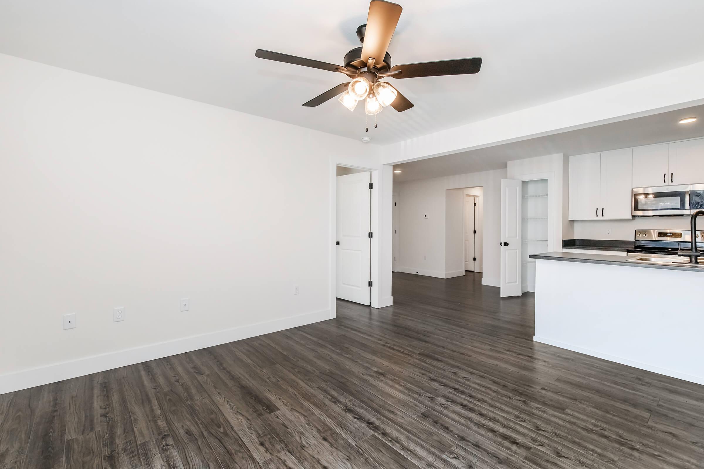 Bright and spacious interior of a modern apartment featuring a ceiling fan, hardwood-like flooring, and white walls. The room includes a doorway leading to another part of the apartment, with an open layout connecting to a kitchen area visible in the background.