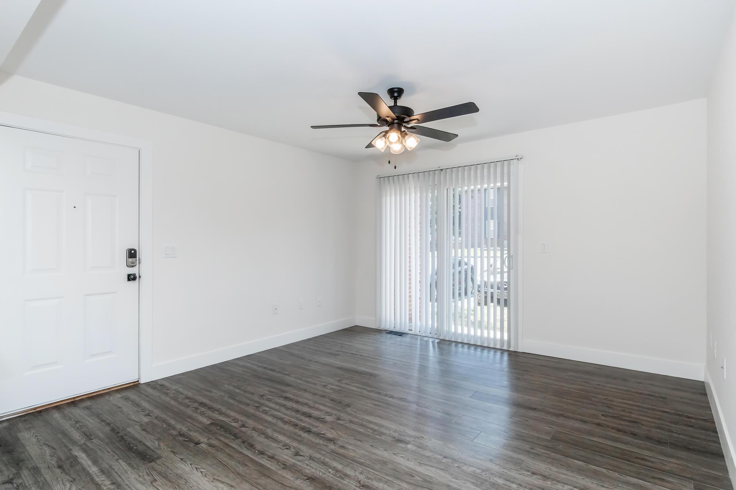 A spacious, empty room with light-colored walls and dark wood flooring. A ceiling fan with five light bulbs is installed at the center. On one side, there's a door leading outside and large sliding glass doors with white blinds, allowing natural light to enter the room.