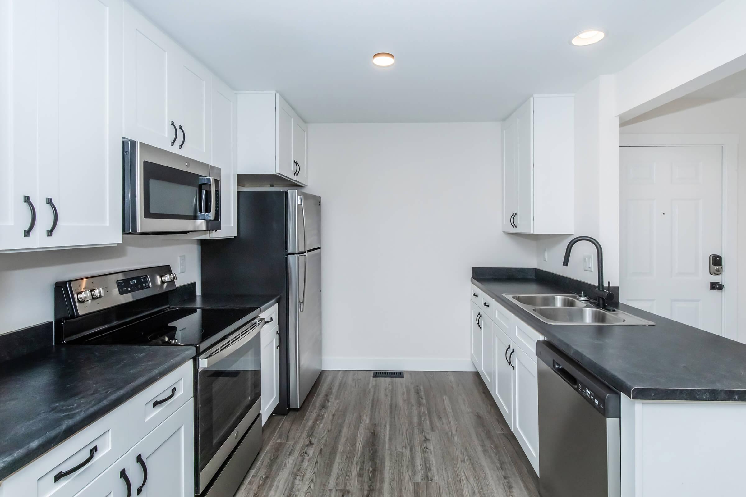 A modern kitchen with white cabinets, stainless steel appliances including an oven, microwave, refrigerator, and dishwasher. The countertops are dark, and the flooring is a light wood finish. The kitchen is well-lit with recessed ceiling lights and features a double sink.