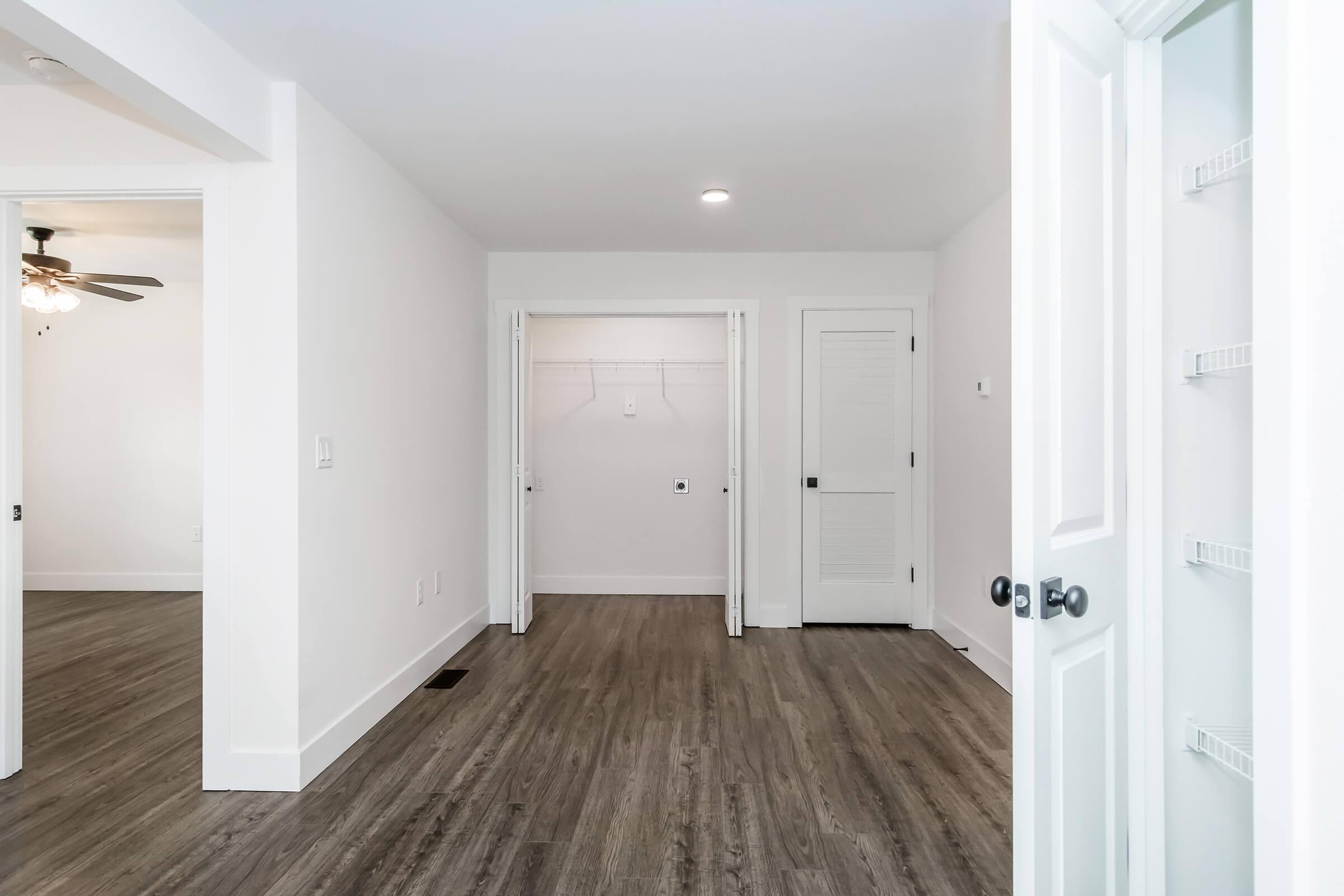 A well-lit, empty room featuring white walls and a smooth, dark wood-like floor. There are open doors leading to closets on the left, with a ceiling fan visible in the background. The space is uncluttered and has a modern, minimalist design.