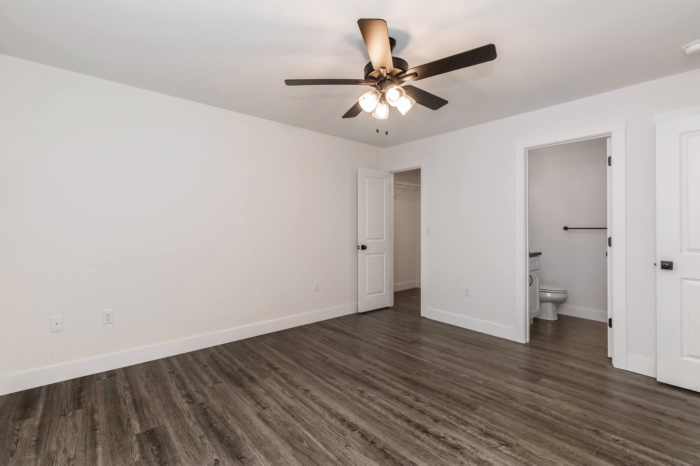 Minimalist room with light-colored walls, a ceiling fan with lights, and dark wood-look flooring. One side features a closed door leading to a closet, and another door leads to a bathroom visible in the distance. The space is bright and uncluttered, perfect for various decorating options.
