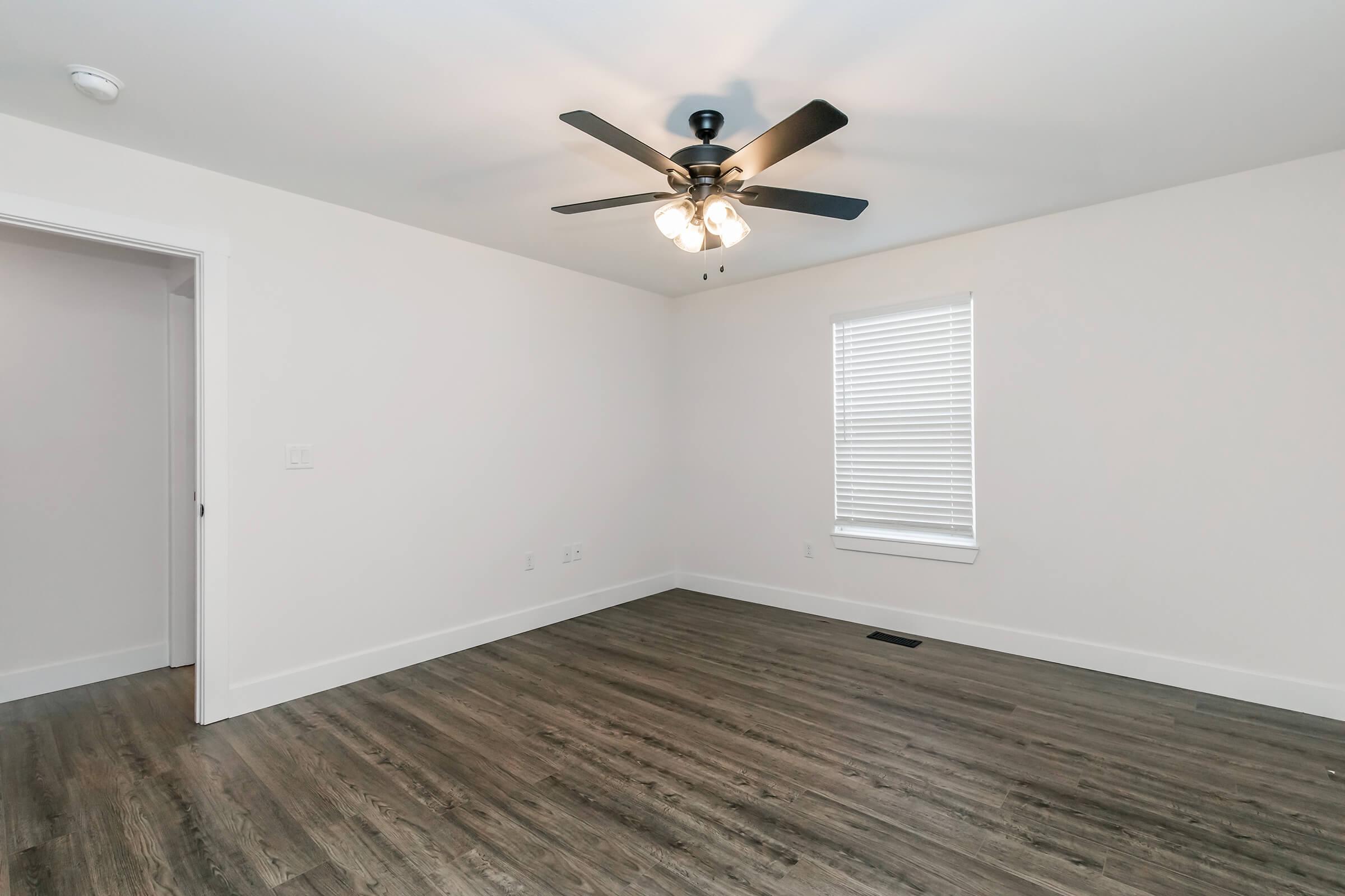 A bright, empty room featuring light-colored walls and a ceiling fan with three bulbs. The floor is a wood-like laminate, and there is a single window with white blinds. A door is partially visible on the left side, adding to the modern, minimalistic aesthetic of the space.