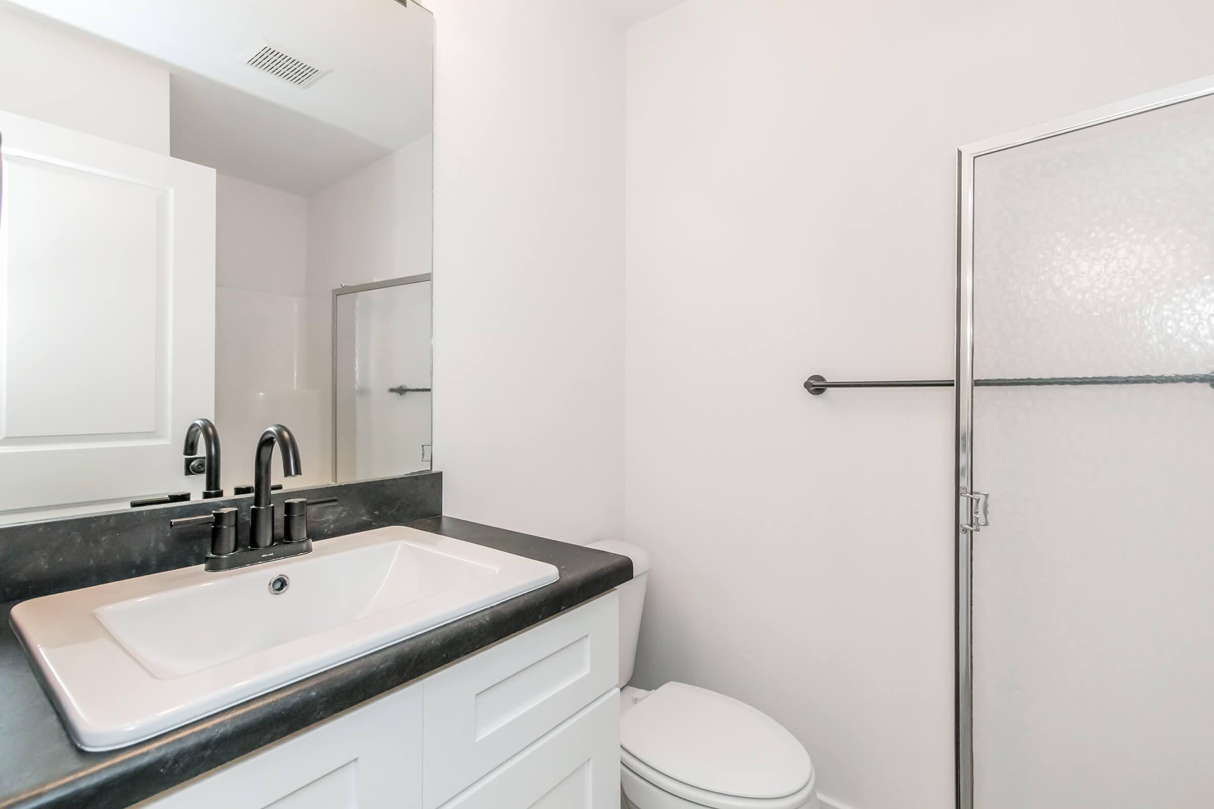 A modern bathroom featuring a rectangular white sink on a black countertop, a sleek faucet, a toilet, and a glass shower enclosure. The walls are painted in a light color, creating a bright and clean atmosphere. A mirror is mounted above the sink.