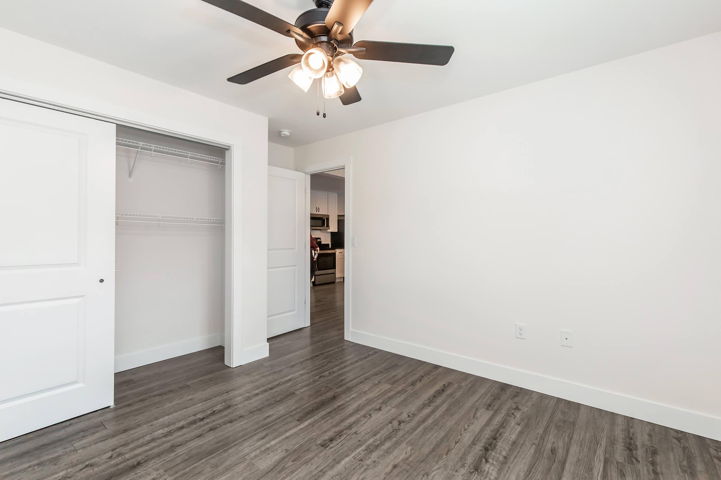 A spacious, empty room with a ceiling fan. One wall features a closed white door and a closet with a hanging rod. The floor is dark wood laminate, and the walls are light-colored. In the background, part of a kitchen can be seen through an open doorway.