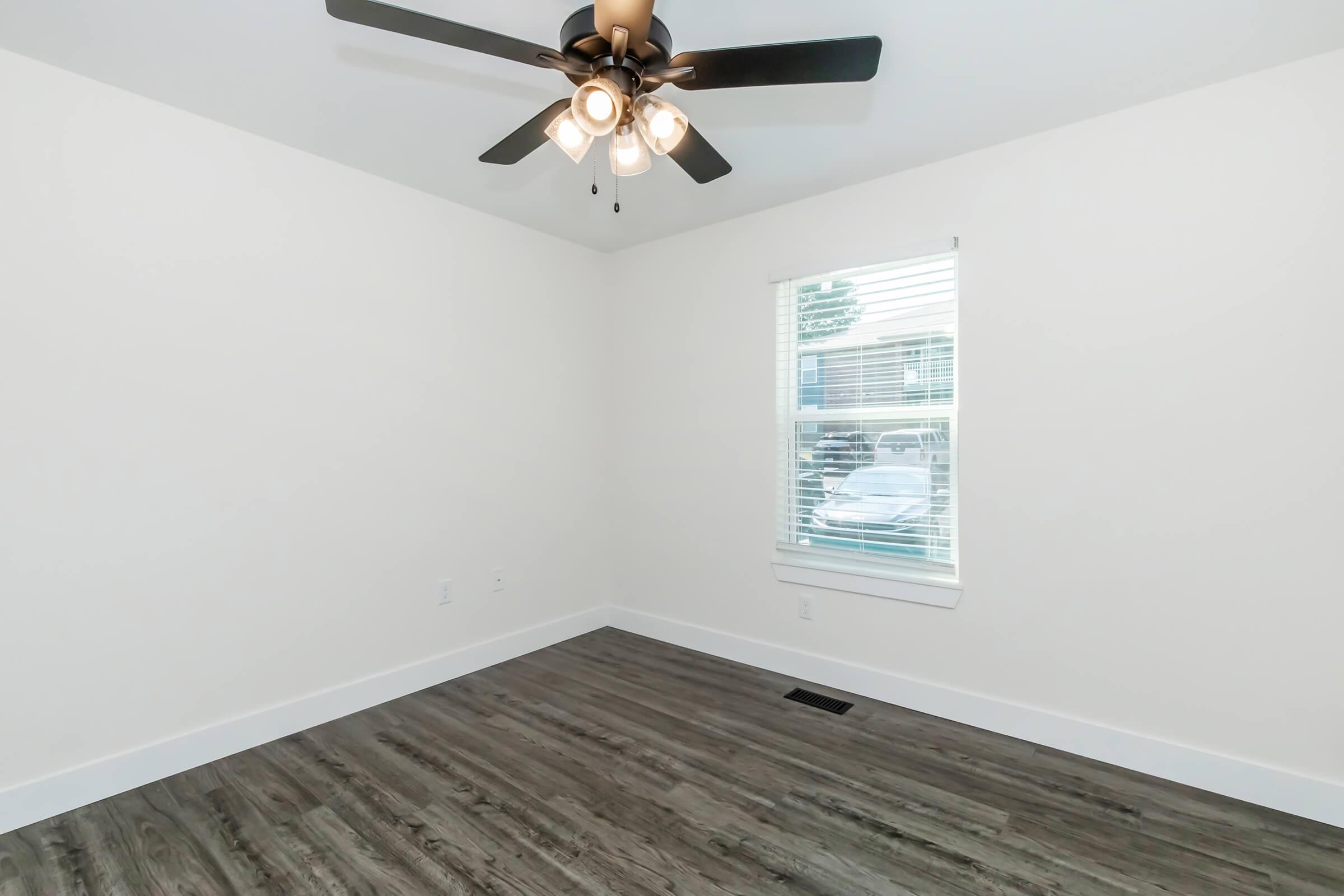 A bright, empty room with a ceiling fan. The walls are painted white, and there is a window with blinds allowing natural light in. The floor features dark, wooden planks, enhancing the modern aesthetic. No furniture is present, making the space feel open and versatile for various uses.