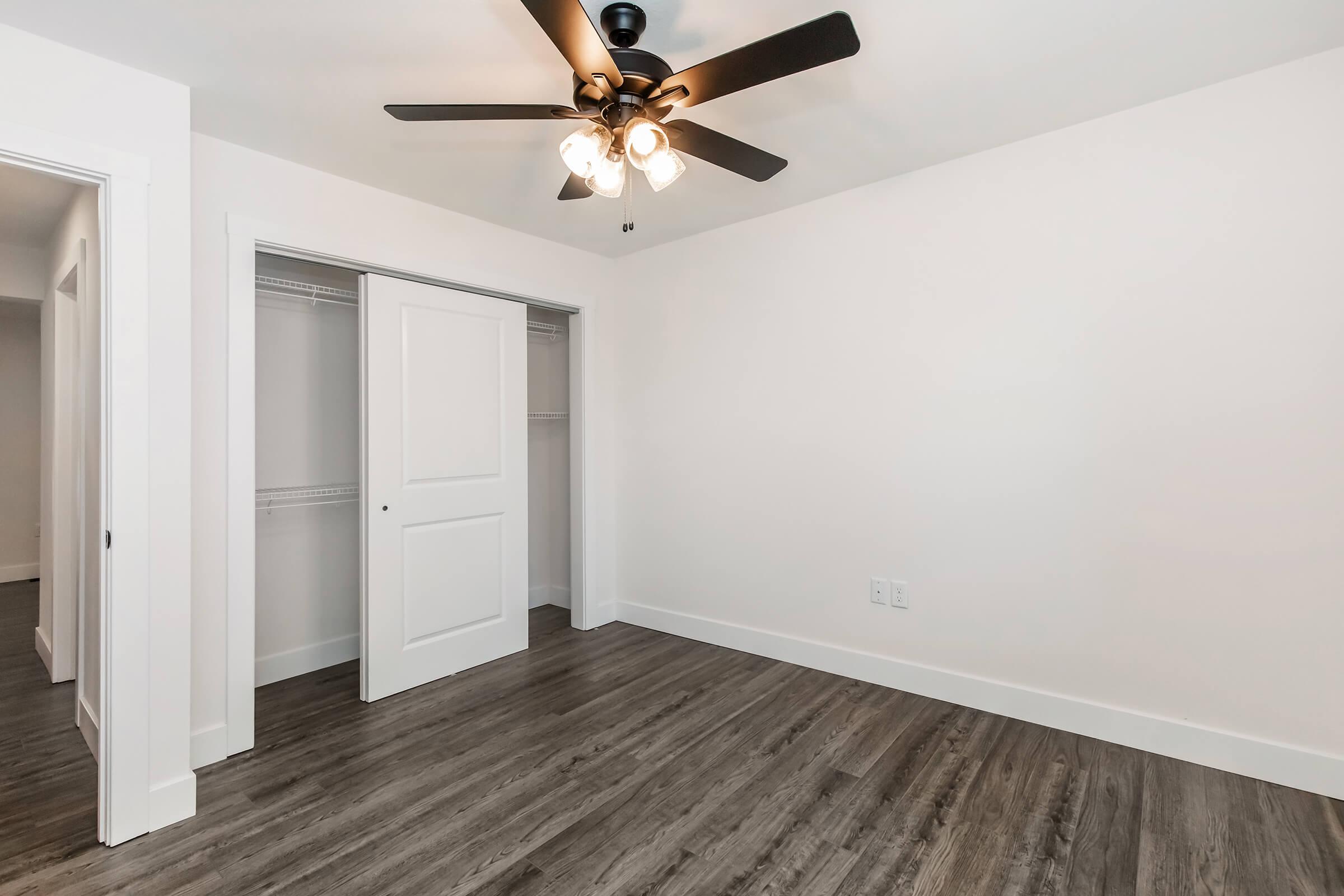 A spacious, minimally decorated room featuring a ceiling fan with light fixtures, light-colored walls, and modern laminate flooring. There is a closed closet with sliding doors on one side, and the room is well-lit with natural light.