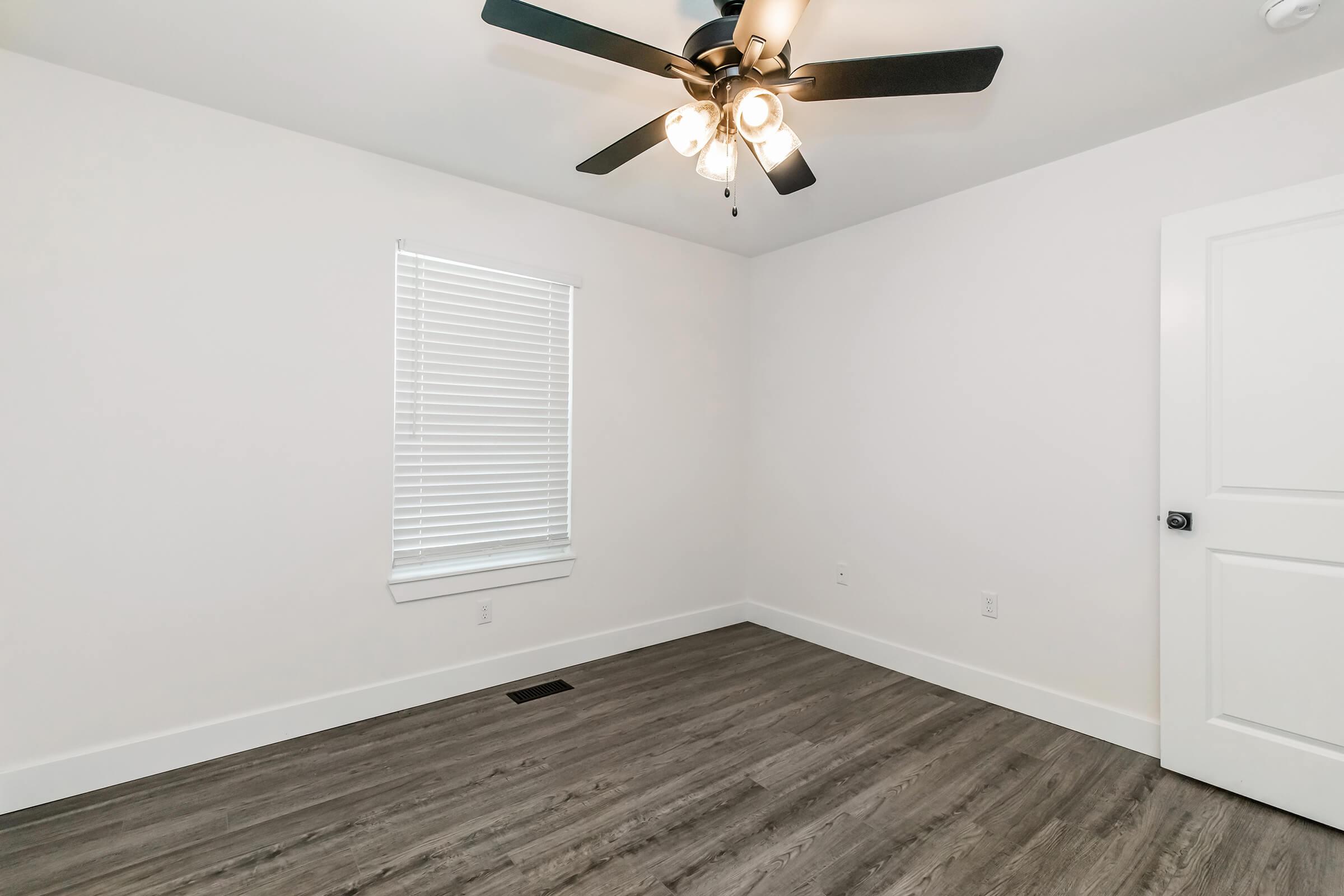 An empty room with light-colored walls and a ceiling fan. The space features a single window with horizontal blinds, allowing natural light. The floor is made of dark, laminate flooring, and there's a white door visible on one side of the room. Overall, the room appears bright and minimally furnished.