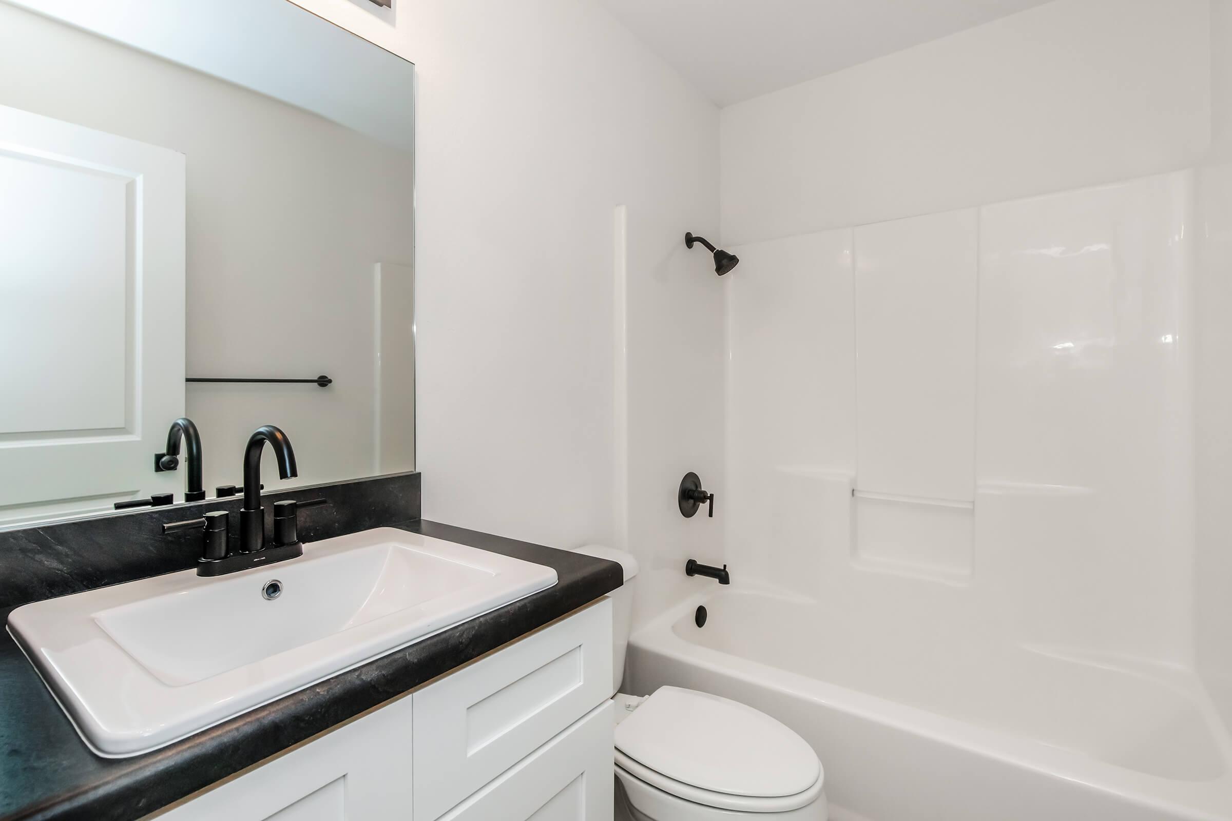 Modern bathroom featuring a white tub and shower combination, a sleek black countertop with a rectangular sink, and elegant black fixtures. A large mirror is mounted on the wall above the sink, and the walls are painted in neutral tones, creating a clean and minimalist aesthetic.