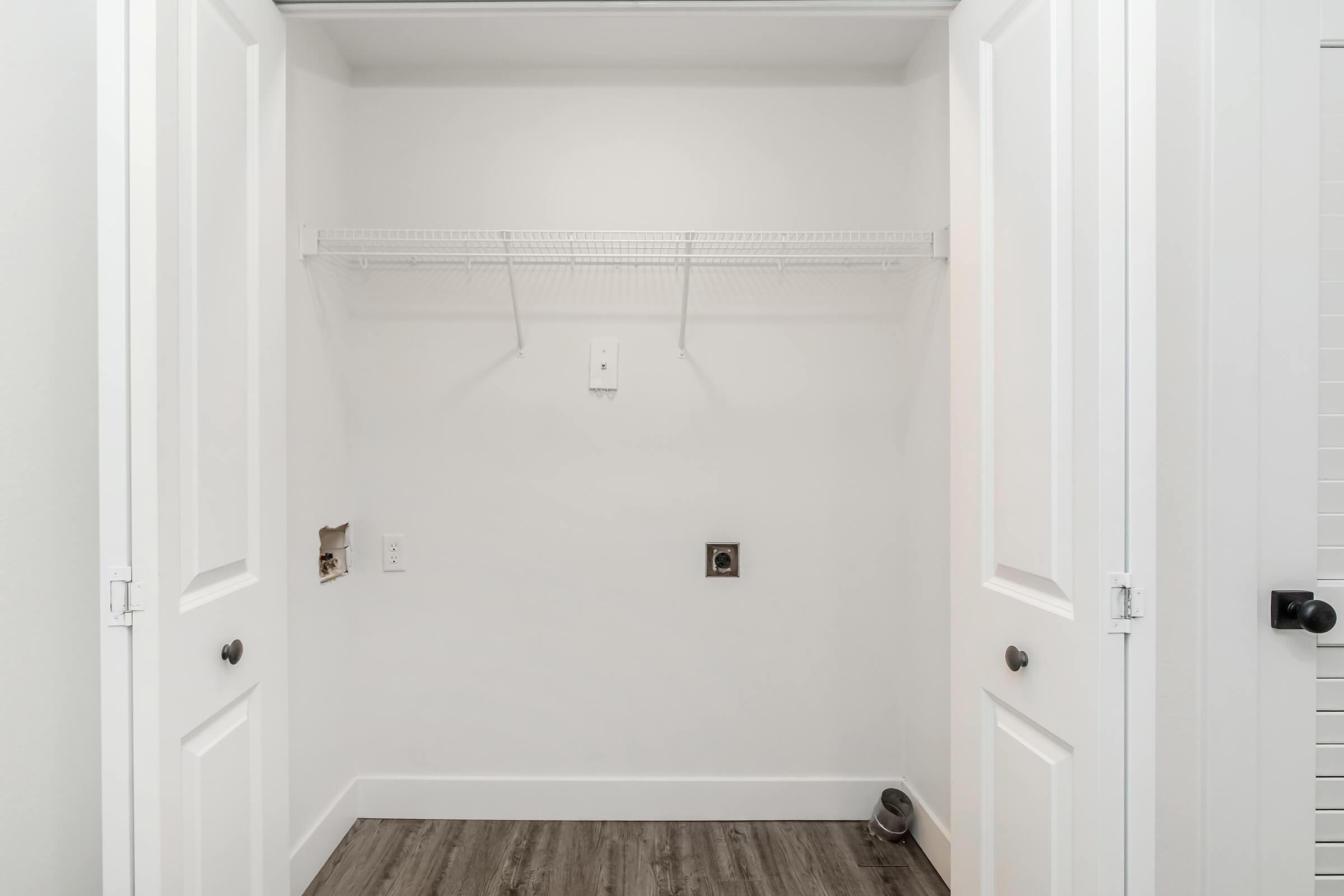 Empty closet with white walls and a wire shelf above. Two white doors are closed, and the floor features light brown wood-like flooring. There is a small wall outlet and vent on one wall, indicating potential for appliances. The space is uncluttered and well-lit, creating a clean, airy appearance.