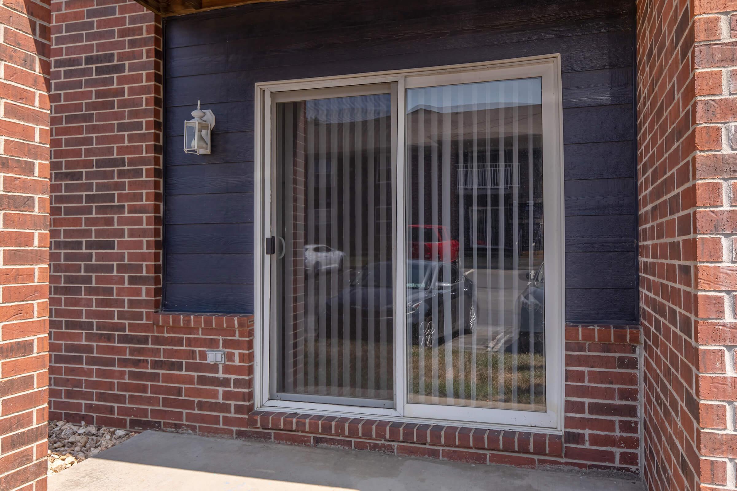 A sliding glass door with vertical blinds, set in a brick exterior wall. The door is flanked by a wall light fixture, and the surrounding area features a concrete patio with some gravel. In the background, a parked vehicle is visible through the glass.