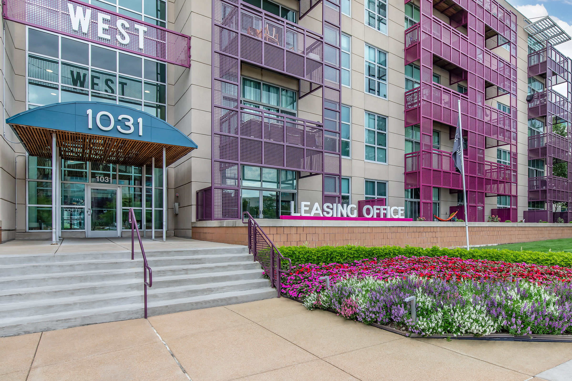 Front view of a modern apartment building with a prominent "WEST" sign. The entrance features a blue awning and stairs leading to the leasing office. Colorful flower beds with various blooms are arranged along the pathway, adding vibrancy to the scene. The building has a contemporary architectural design with large windows.