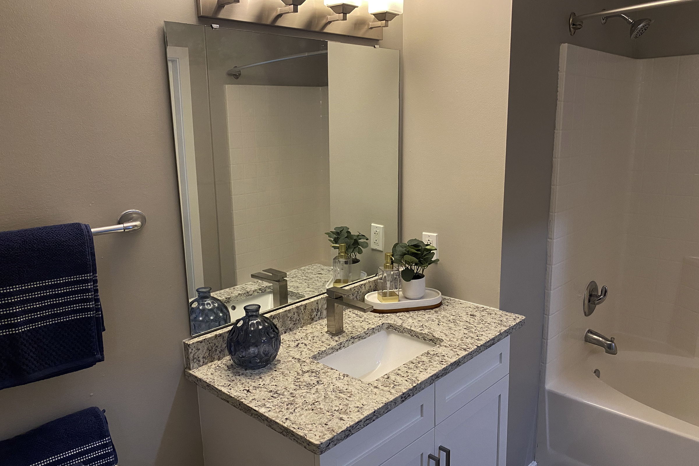 A modern bathroom featuring a white vanity with a granite countertop, a large mirror, and a chrome faucet. A small potted plant and decorative items are placed on the counter. A blue towel hangs on a rack nearby, and a white bathtub with a shower curtain is visible in the background. Soft lighting enhances the serene atmosphere.