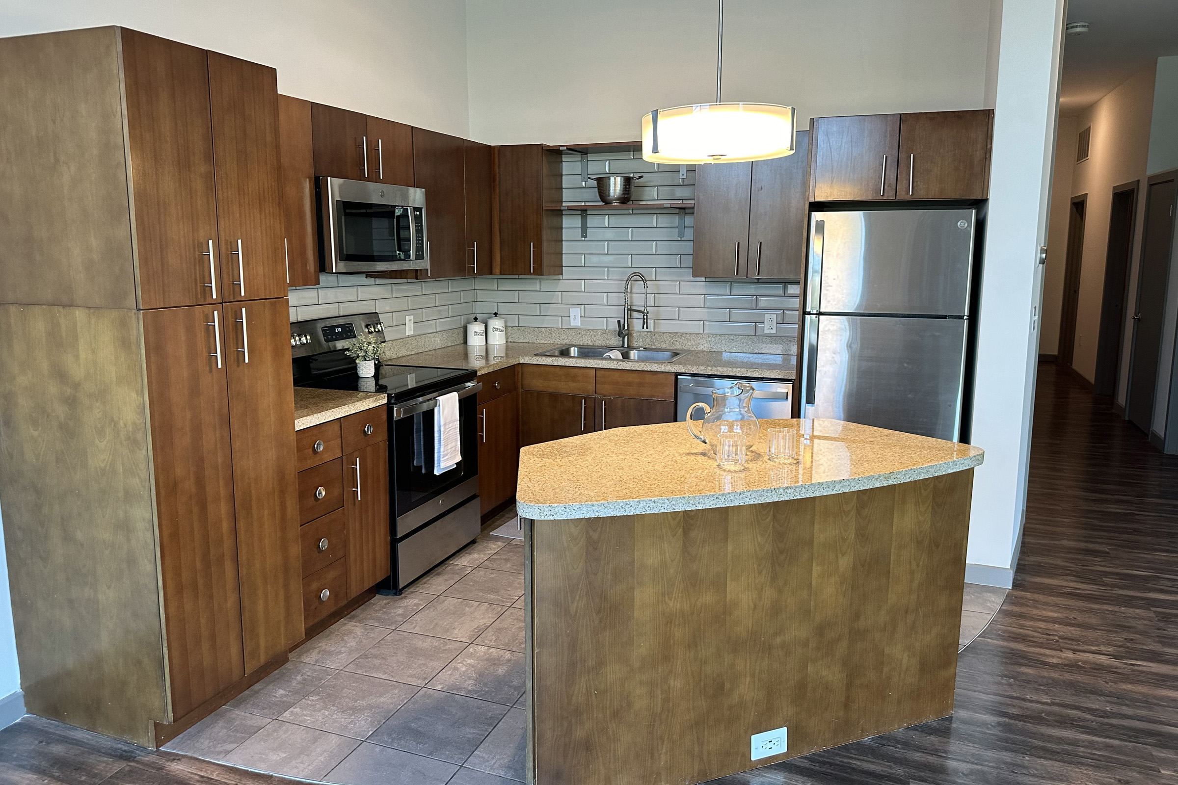Modern kitchen with wooden cabinets, stainless steel appliances, and granite countertops. There is a large island with a curved edge and a few glass items on it. The backsplash consists of gray tiles, and there is a pendant light hanging from the ceiling. The flooring is dark, enhancing the contemporary look.