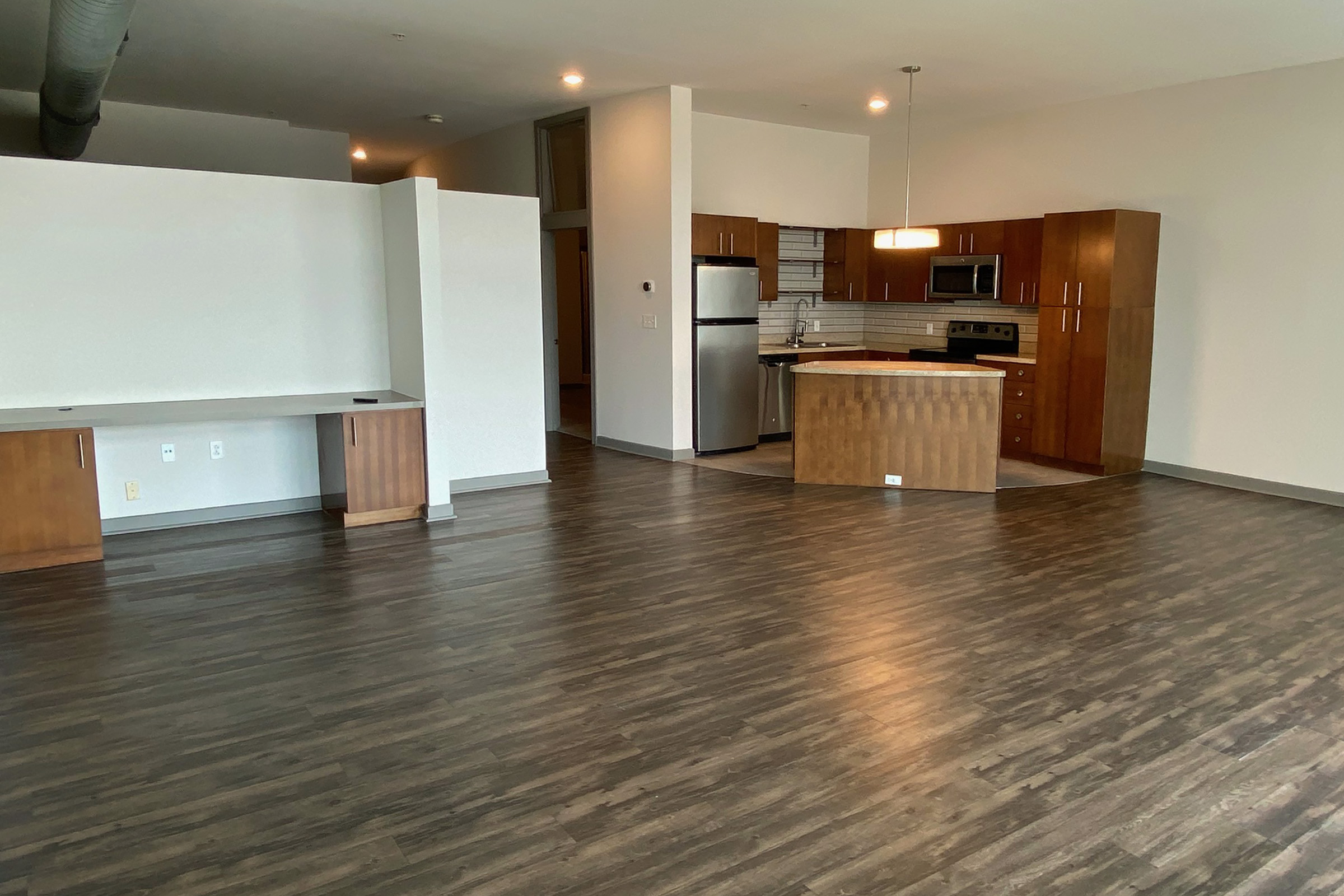 Spacious modern apartment interior featuring an open layout with hardwood floors, a kitchen equipped with stainless steel appliances, wooden cabinetry, and a central island. Natural light floods the room, highlighting the clean design and neutral color palette.
