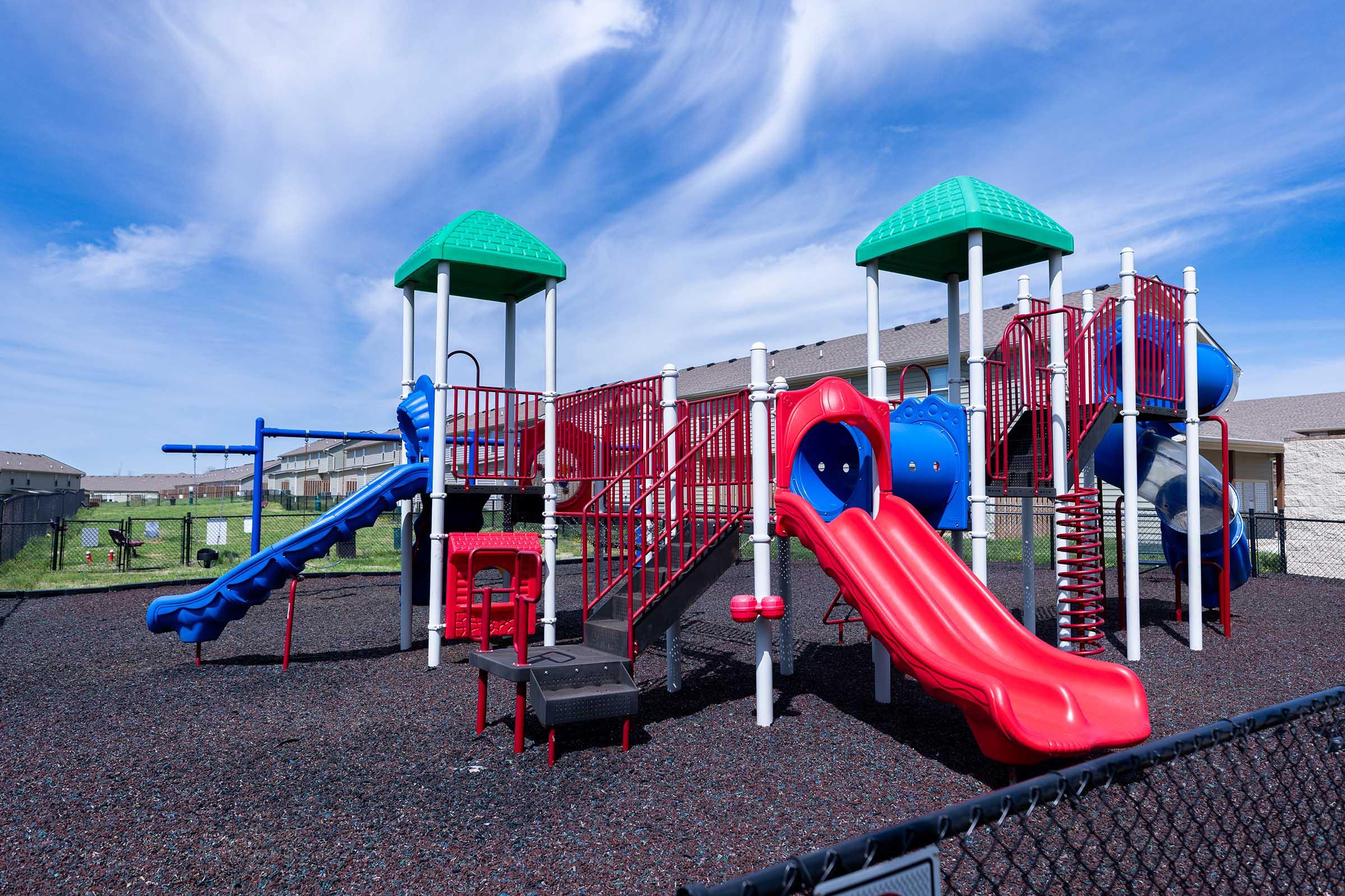 Colorful children's playground featuring blue and red slides, climbing structures, and safety features. It is surrounded by a fence and set against a backdrop of a blue sky with wispy clouds. Grass and buildings are visible in the background, creating a vibrant play area for kids.