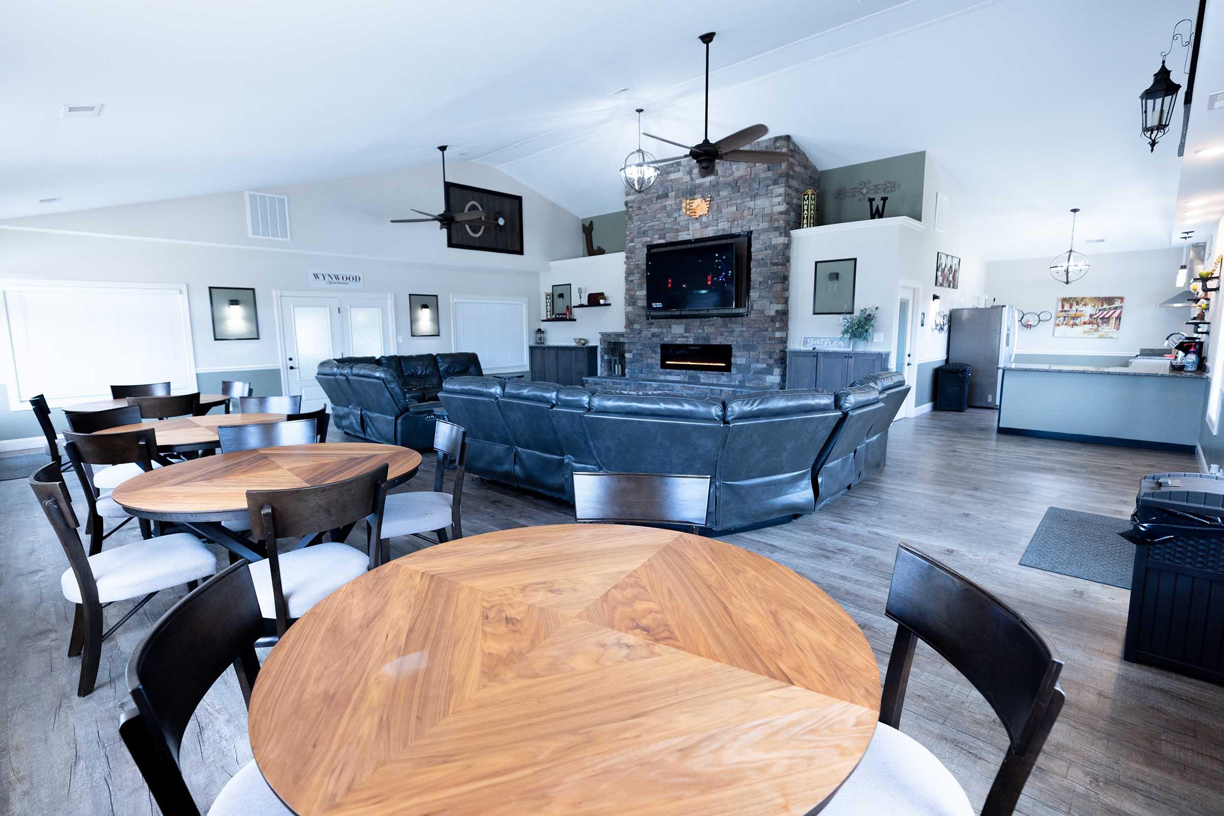 A spacious, modern living area featuring a large sectional sofa facing a stone fireplace with a mounted TV above. Wooden dining tables with black chairs are visible in the foreground. The bright space has large windows, stylish ceiling fans, and a clean, contemporary design.