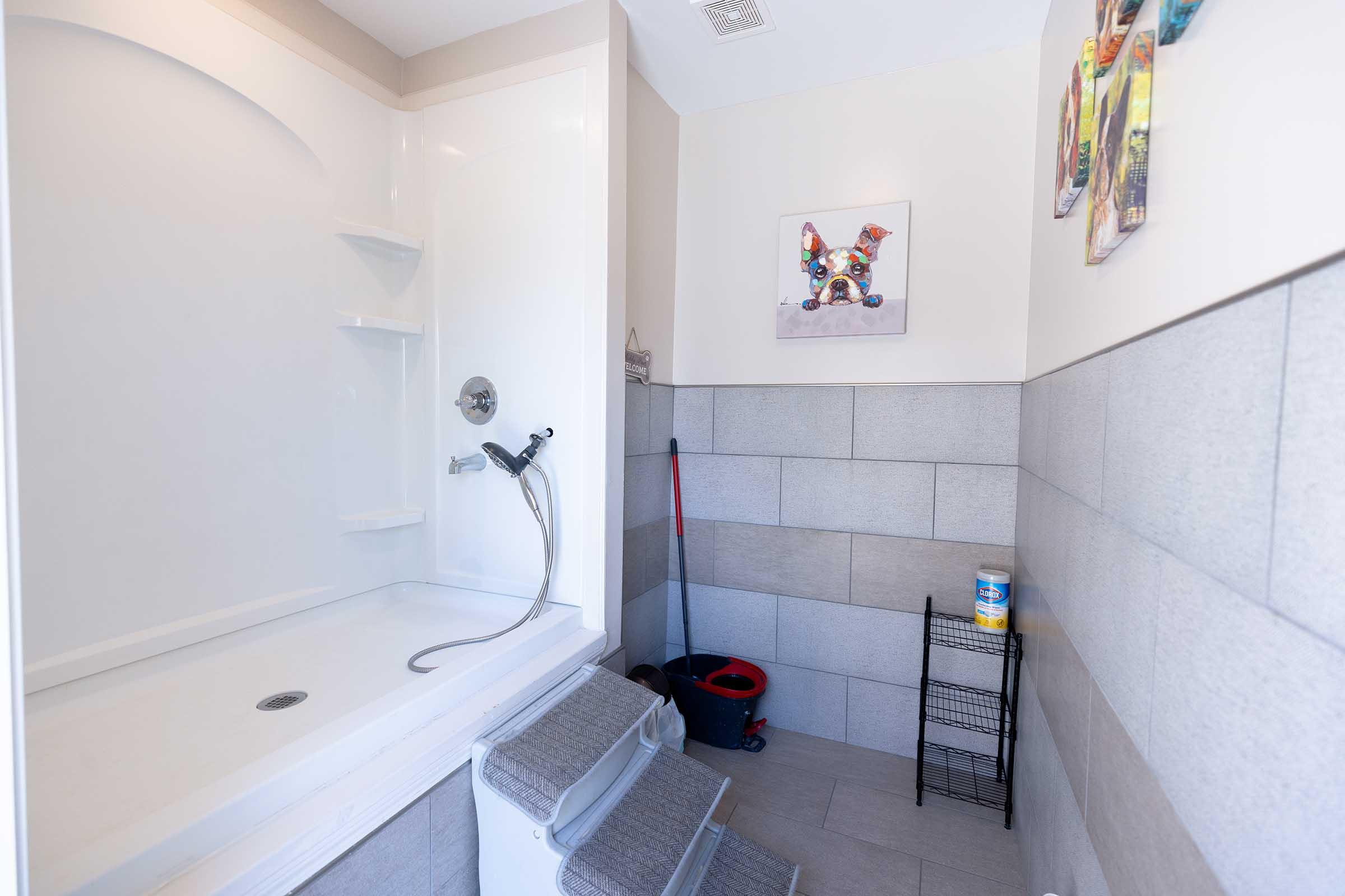 A modern bathroom featuring a white shower stall with a handheld showerhead, gray tile walls, and a light-colored floor. A small bench is placed outside the shower, along with cleaning supplies in a black wire basket and a mop. Wall art is visible, adding a decorative touch to the space.
