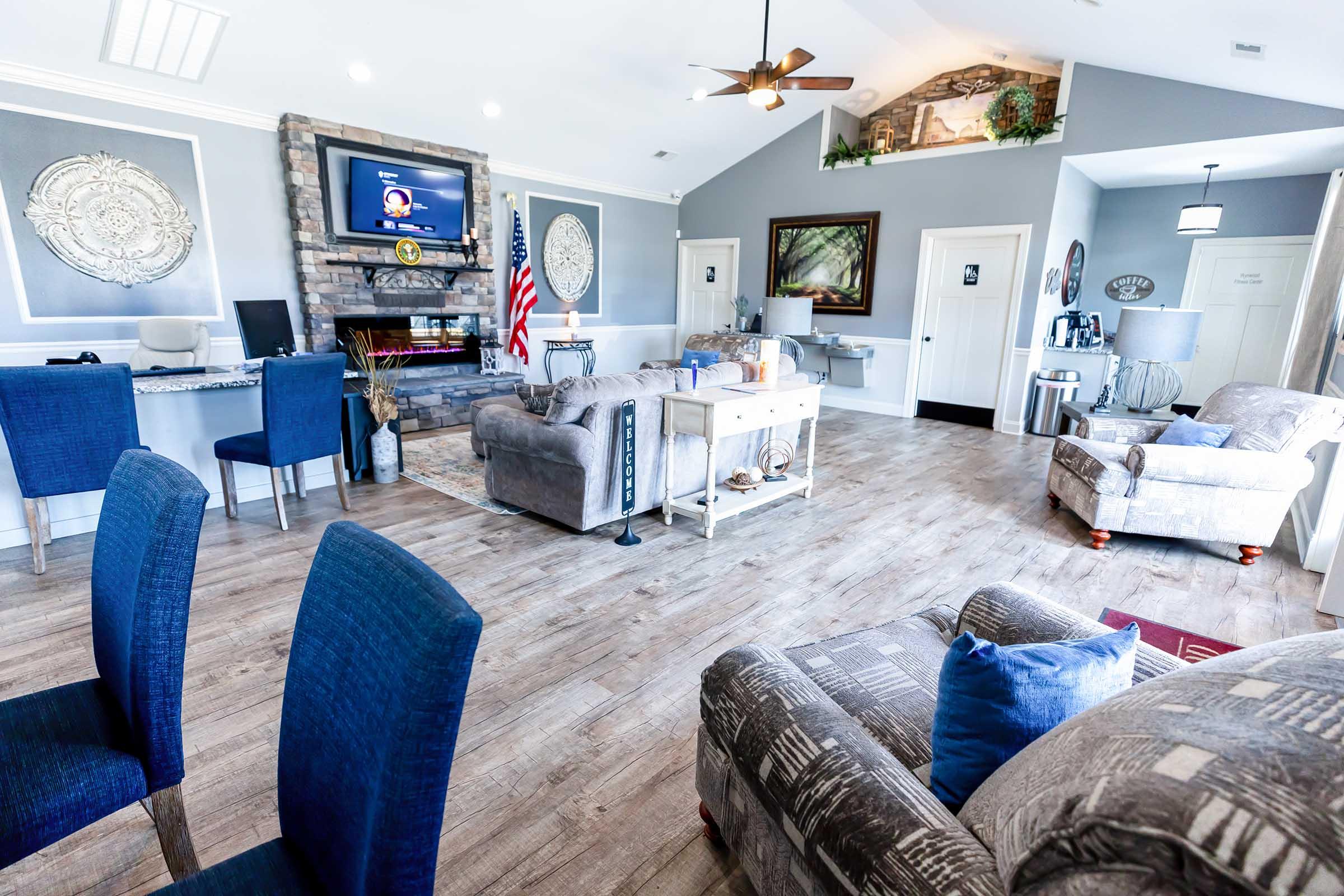 A spacious living area featuring modern furniture, including a couch, chairs, and a coffee table. There’s a stone fireplace with a TV mounted above it, and an American flag displayed. The walls are painted in a neutral color, with decorations and artwork. Natural light fills the room from windows and a ceiling fan.
