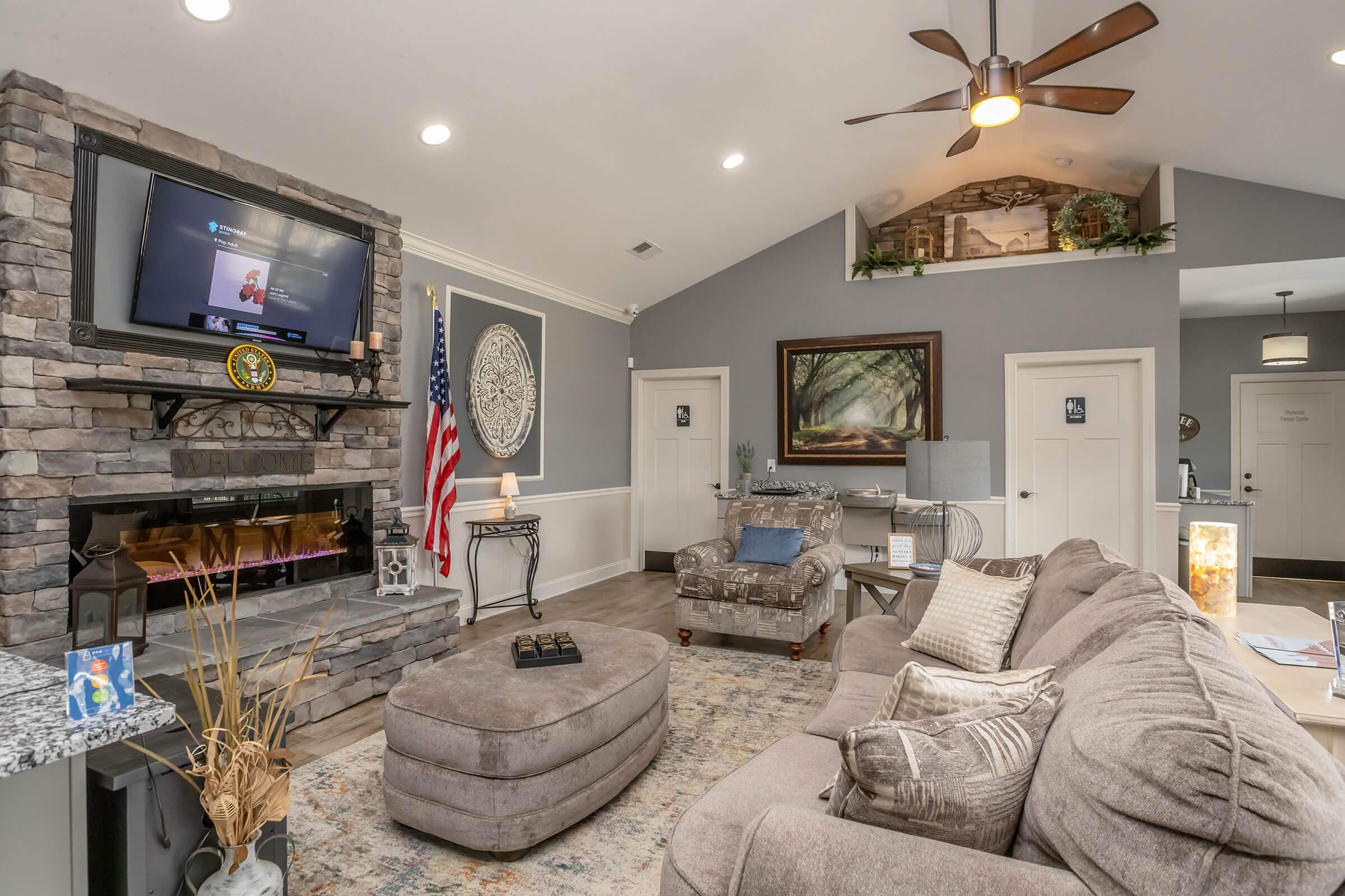 Cozy living room featuring a stone fireplace with a TV above, an American flag, and a large sectional sofa. Light gray walls and decorative accents create a warm atmosphere. A potted plant adds a touch of greenery, while a coffee table sits in front of the sofa, enhancing the inviting space.