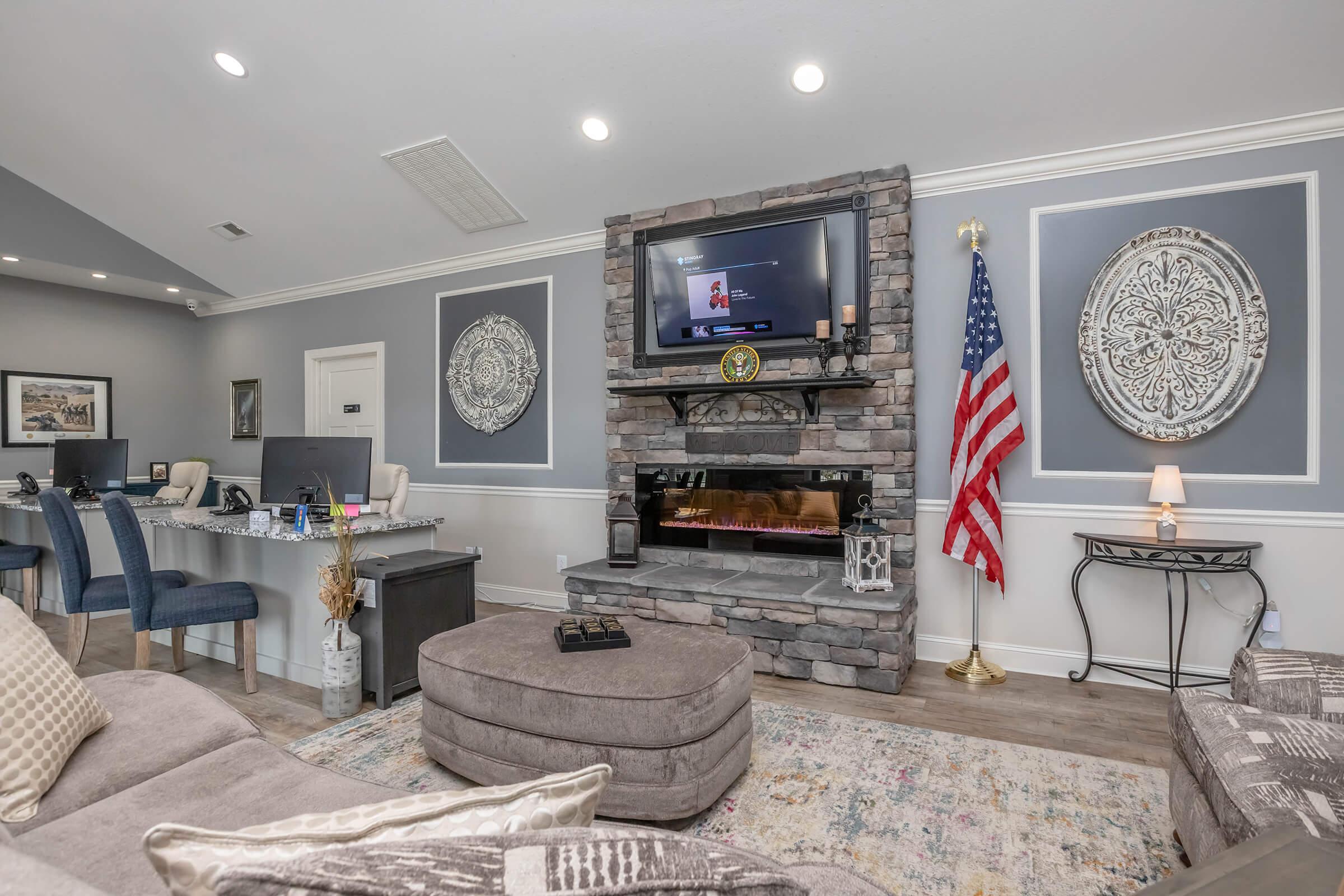 A cozy living room featuring a stone fireplace with an electric insert, a mounted TV above, and decorative wall accents. There's a plush gray sofa and a colorful area rug. An American flag is displayed, and a small table with a lamp adds to the inviting atmosphere.