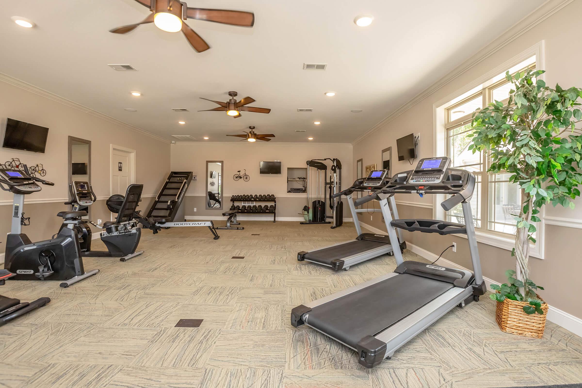 A modern fitness gym featuring treadmills, stationary bikes, strength training equipment, and wall-mounted TVs. The space is well-lit with ceiling fans and large windows, creating an inviting atmosphere for workouts. potted plants add a touch of greenery to the environment.