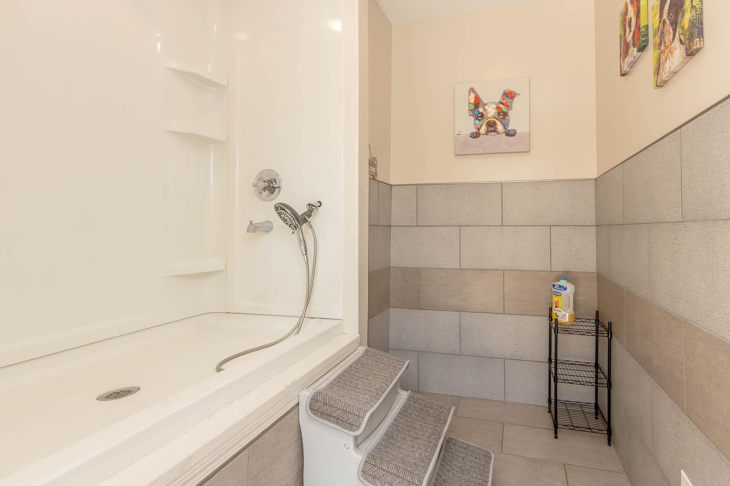 A clean bathroom featuring a shower with a handheld showerhead, grey tiled walls, and light-colored decor. There is a small step stool for accessibility and a metal rack holding toiletries. A colorful artwork is hung on the wall above the shower area, adding a decorative touch to the space.