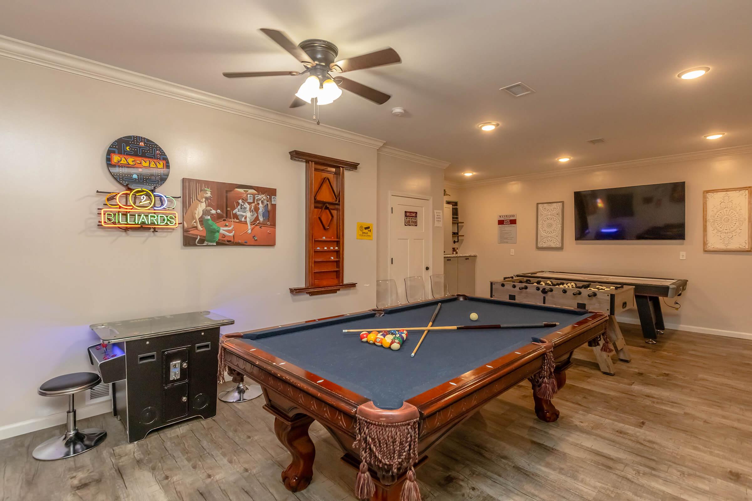 A cozy game room featuring a stylish billiards table with pool cues, a foosball table, a wall-mounted TV, and various decorations, including neon signs and framed artwork. Warm lighting and wooden flooring create an inviting atmosphere for gaming and entertainment.